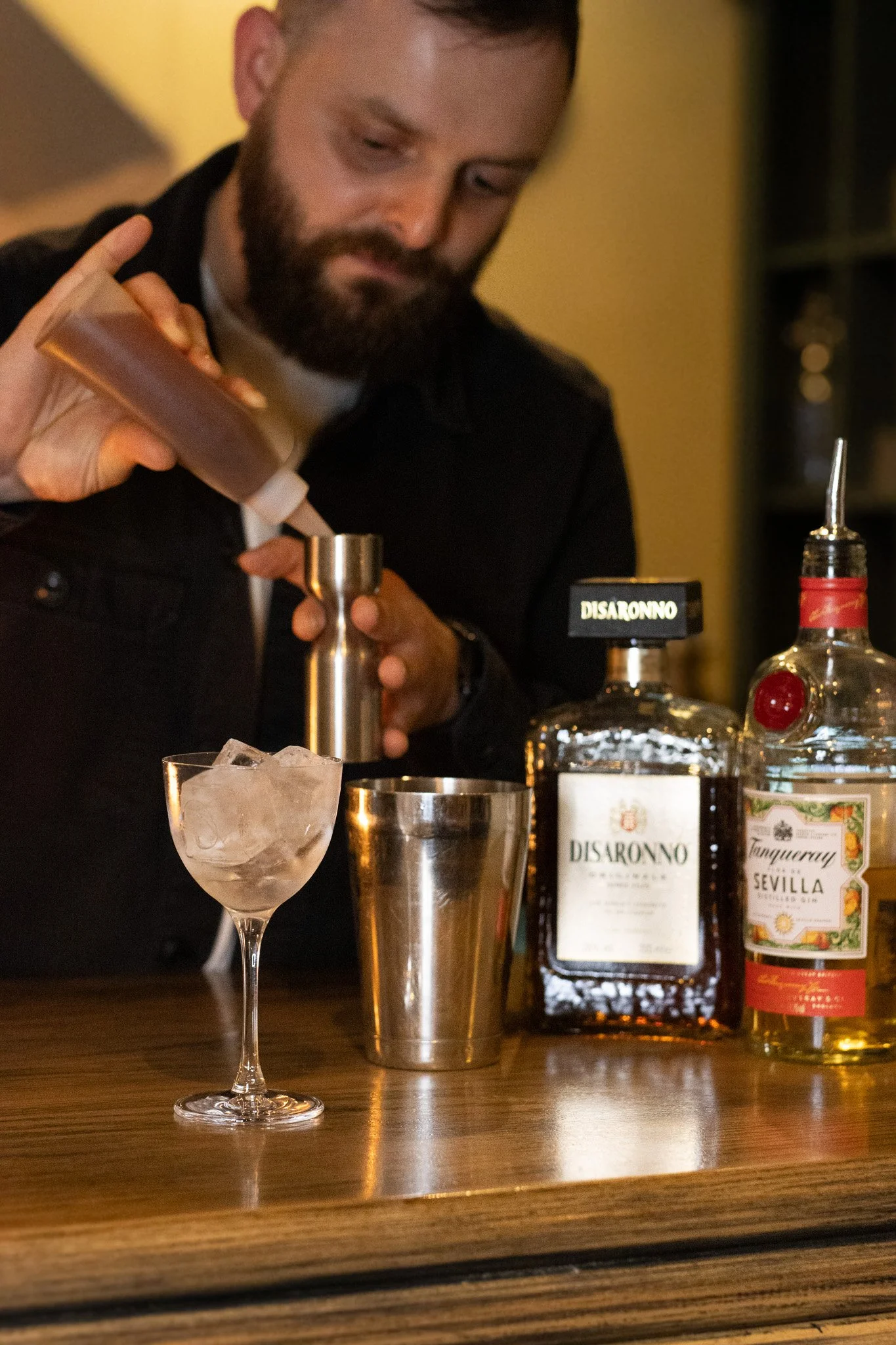 A bartender preparing a cocktail behind a bar. The image shows a glass with ice, bottles of alcohol including Disaronno and Tanqueray gin, and bar tools. The Lamb Inn. 