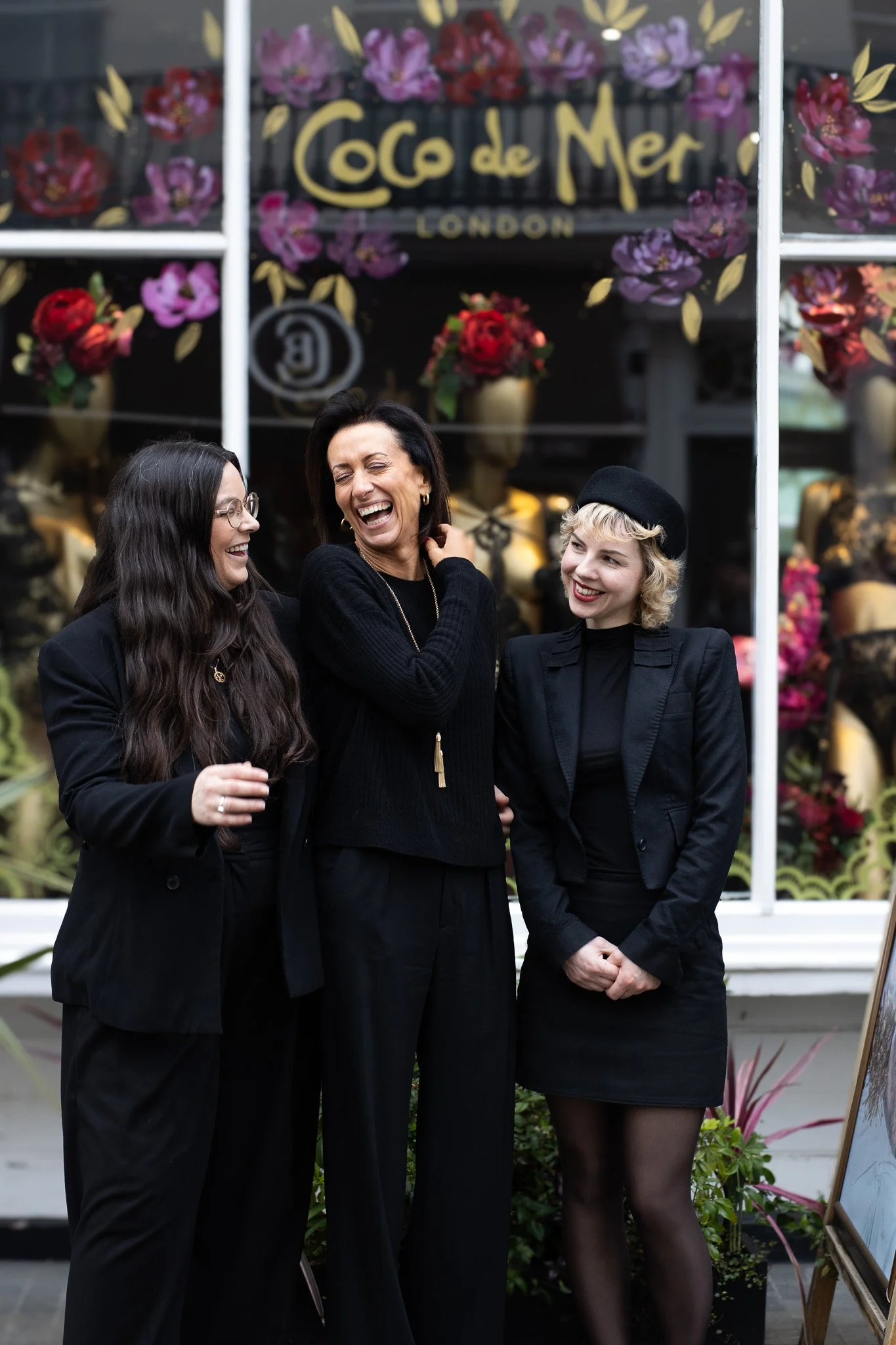 Three women are standing together in front of a floral shop window that has the sign 'Coco de Mer London'. They are dressed in black, smiling, and appear to be having a joyful conversation. Coco De Mer.