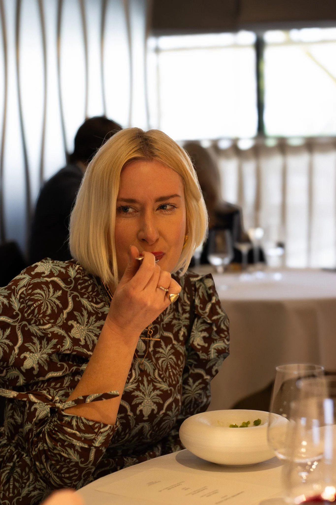 A woman with blonde hair sitting at a dining table, looking to her left, with a thoughtful expression. In front of her is a white bowl of soup garnished with herbs, and there are wine glasses on the table. Petrus.