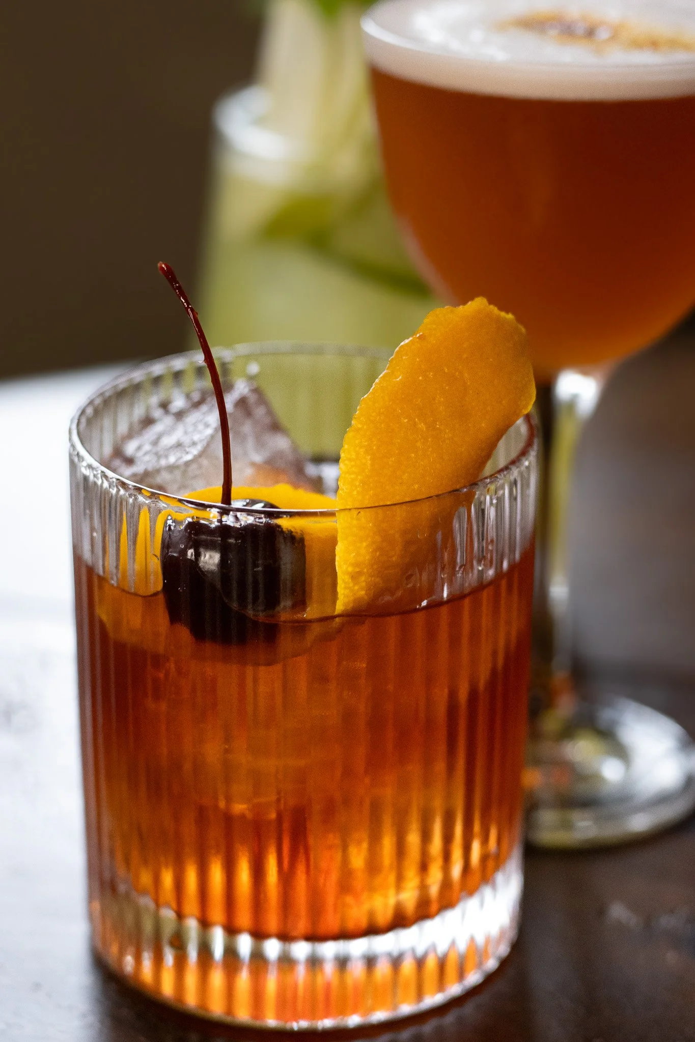 Close-up of a cocktail in a glass with orange slice garnish, a maraschino cherry on top, and a large beer in a glass in the background. The Lamb Inn. 