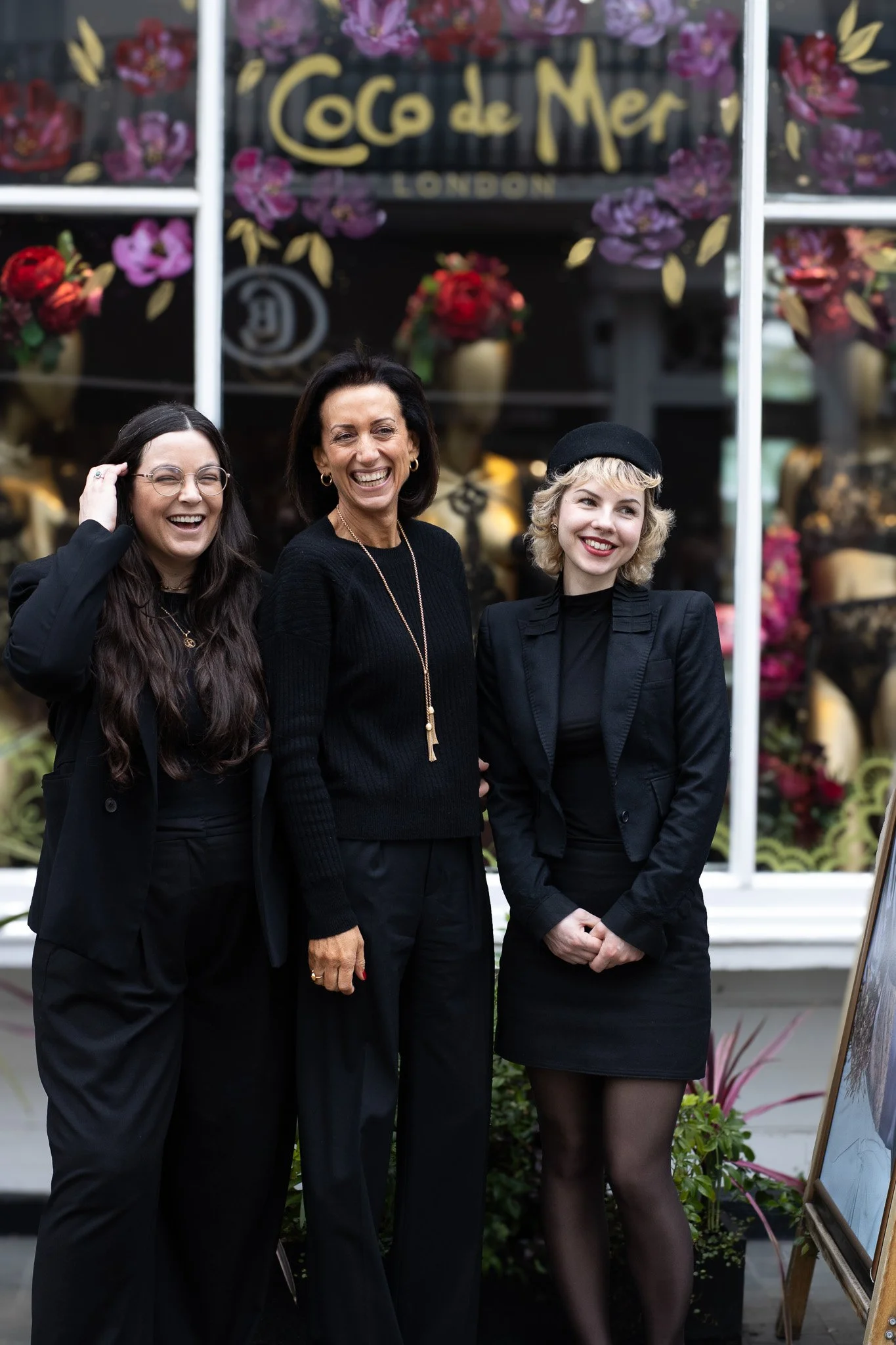 Three women dressed in black professional attire standing outdoors in front of a flower shop window, smiling for a photo. Coco De Mer.