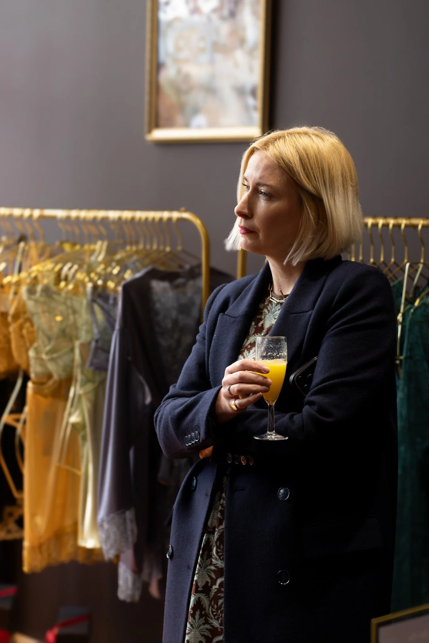 A woman with blonde hair wearing a dark blazer, holding a glass of orange juice, standing in front of clothing racks and a mirror. Coco De Mer.