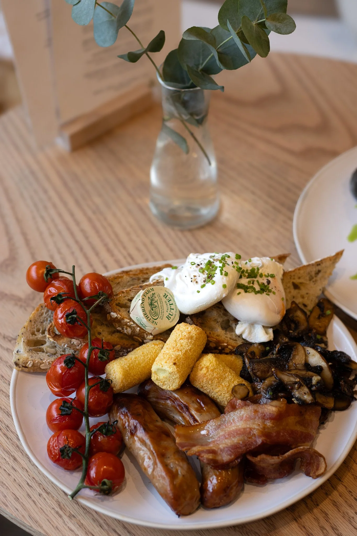 Breakfast plate with cherry tomatoes, bacon, mushrooms, eggs, potatoes, bread, and a sprig of cherry tomatoes on the side, on a white plate. The Cotswold Farm. 