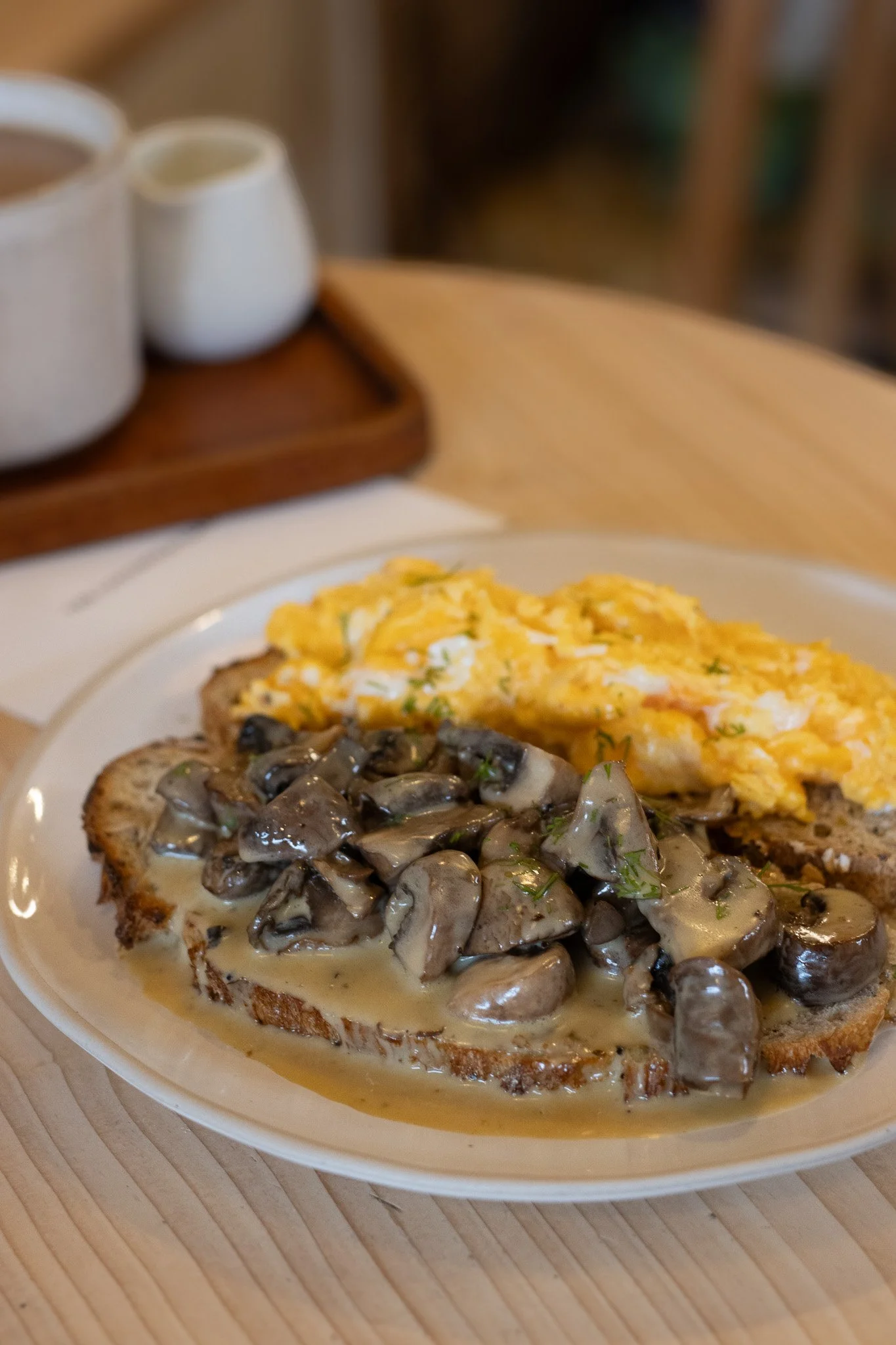Close-up of avocado toast topped with sautéed mushrooms, scrambled eggs, and shredded cheese, served on a white plate on a wooden table. The Cotswold Farm. 