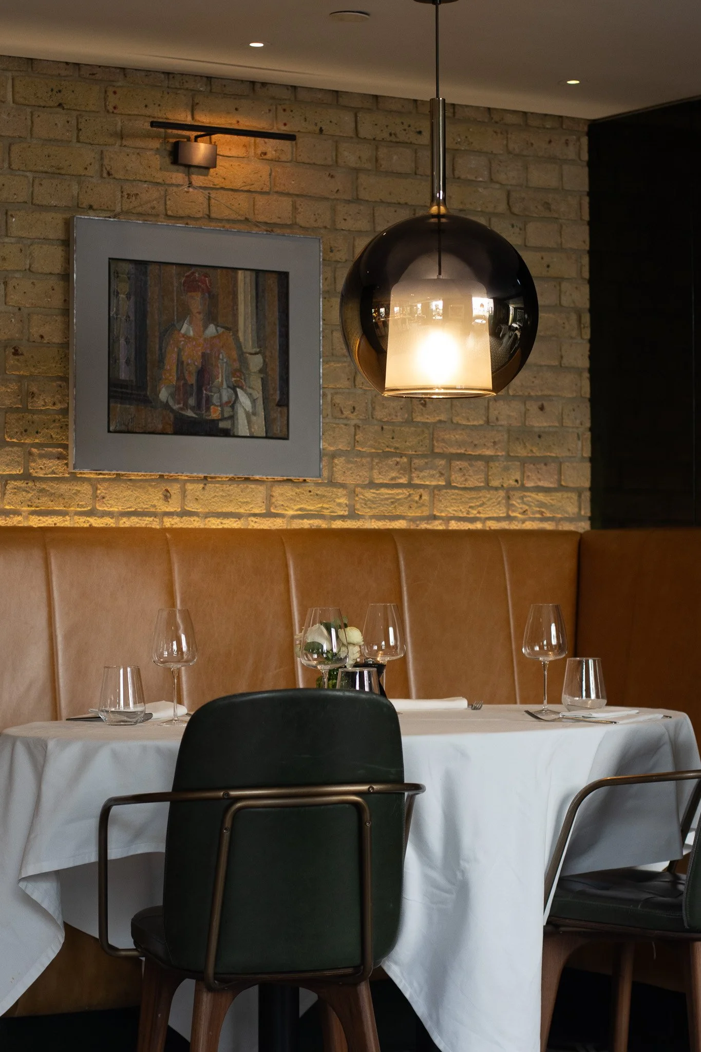 A restaurant dining area with a round table covered with a white tablecloth, set with wine glasses, a small flower arrangement, and silverware. The Hari, Il Pampero.