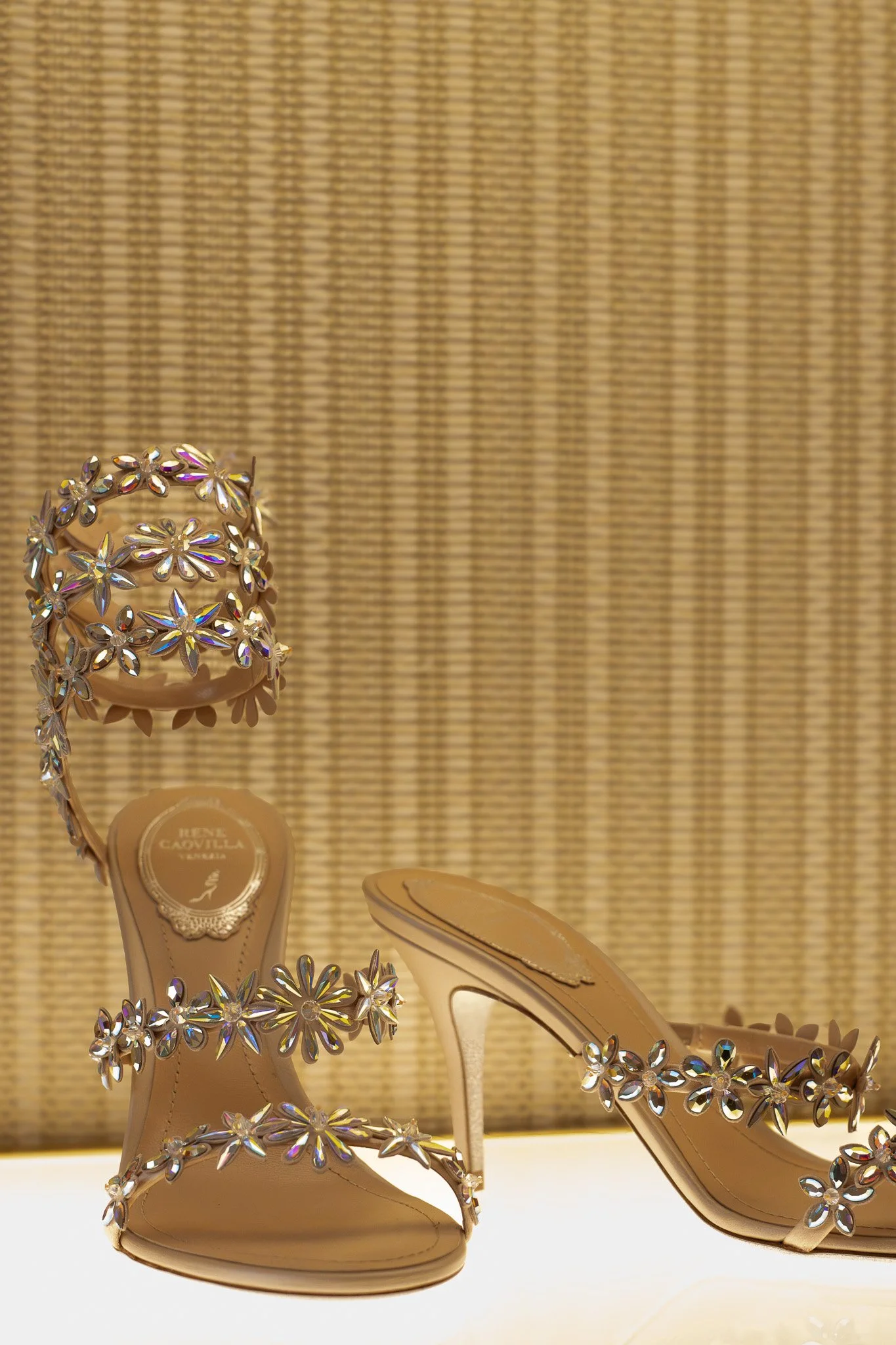 Beige high-heeled sandals decorated with flower-shaped rhinestones, displayed on a white surface with a textured golden background. Rene Caovilla.