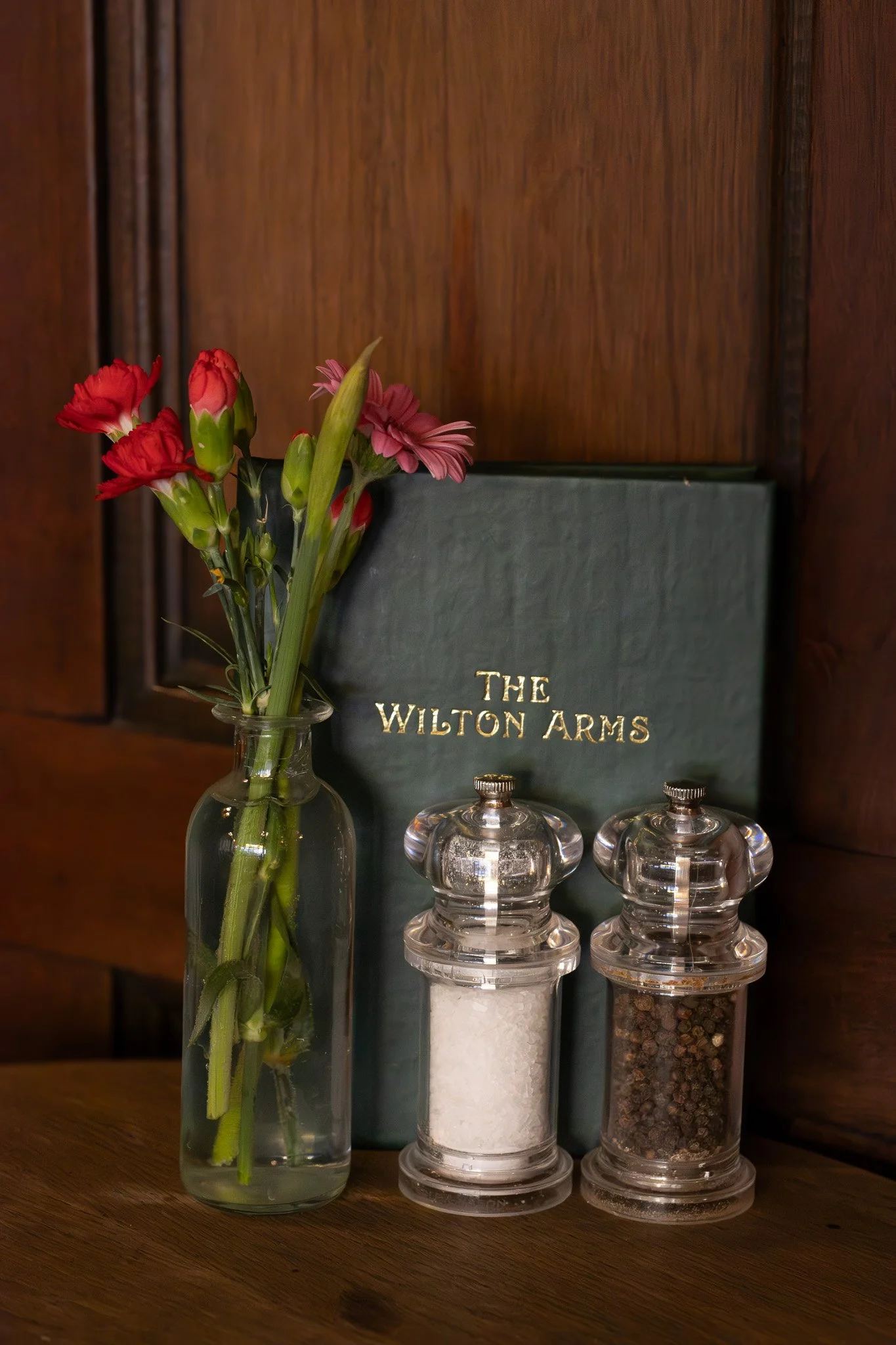 A vase of red and pink flowers, a green book titled 'The Wilton Arms', and three glass spice jars with white, black, and mixed spices on a wooden surface.  The Wilton Arms.