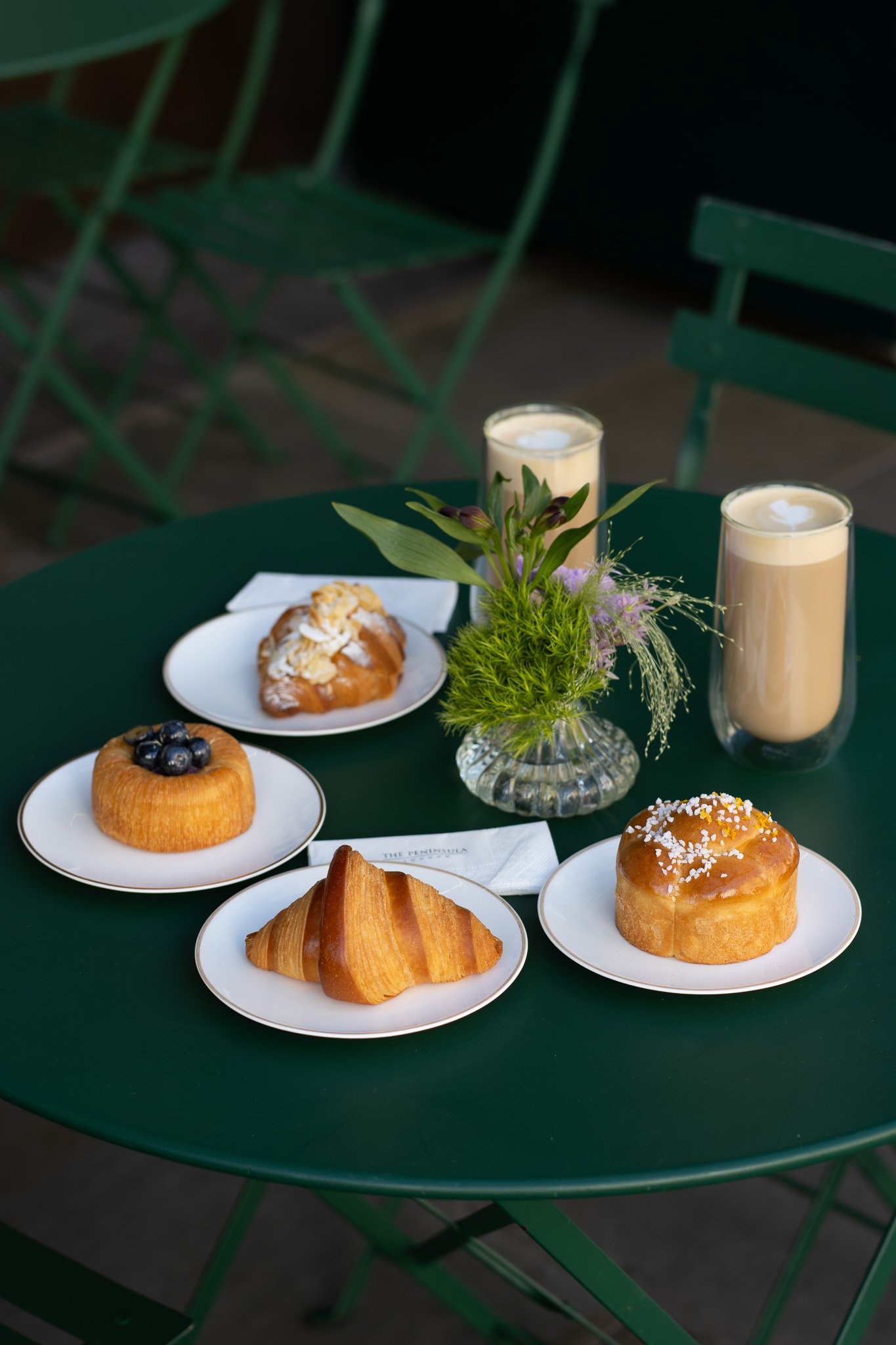 A green round table with assorted pastries, two tall glasses of creamy beverages, and a small floral arrangement in a glass vase. The Peninsula.