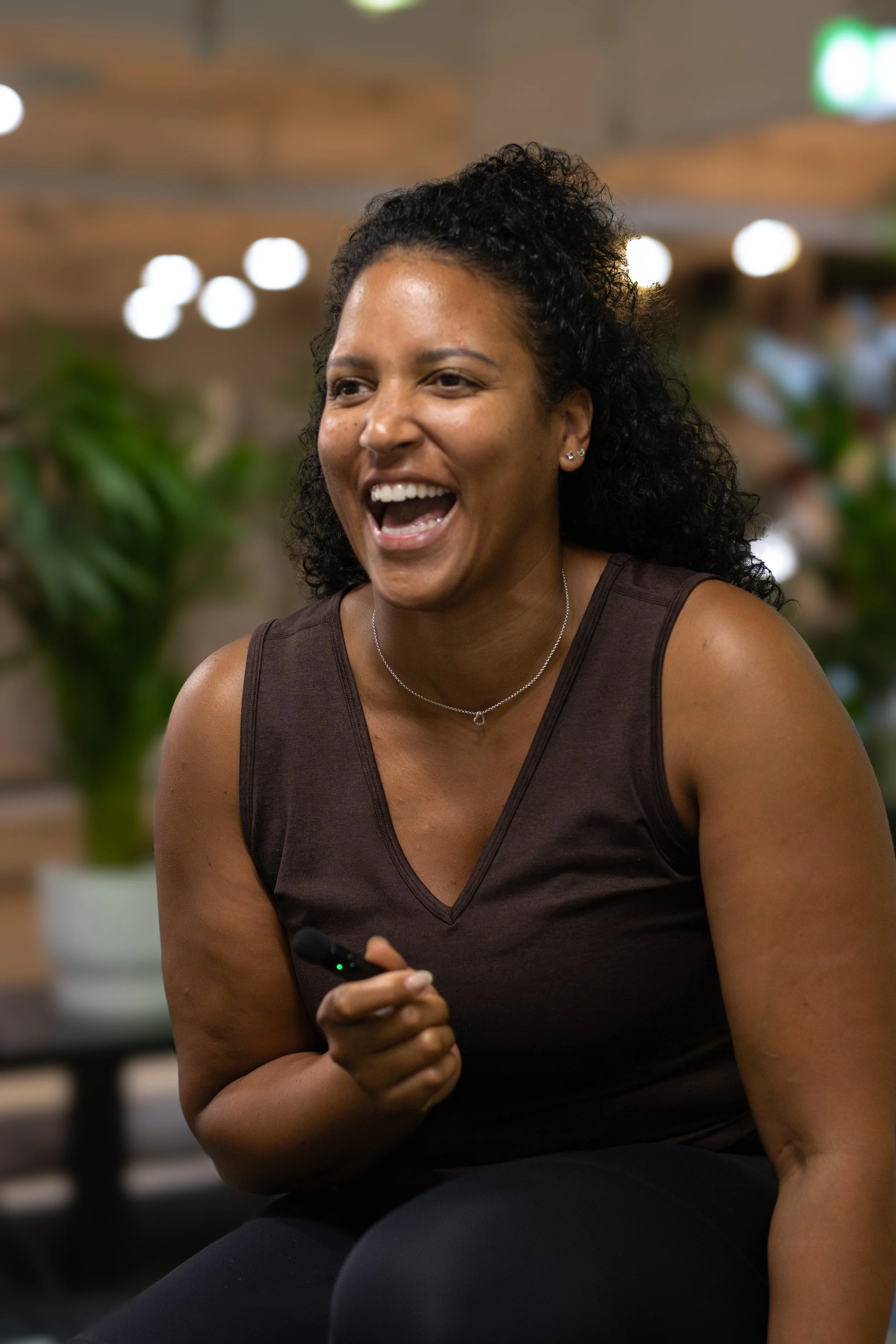 A woman smiling and holding a remote control in her hand, sitting indoors with blurred lights and plants in the background. The Studio Cheltenham, Reformer Pilates. 