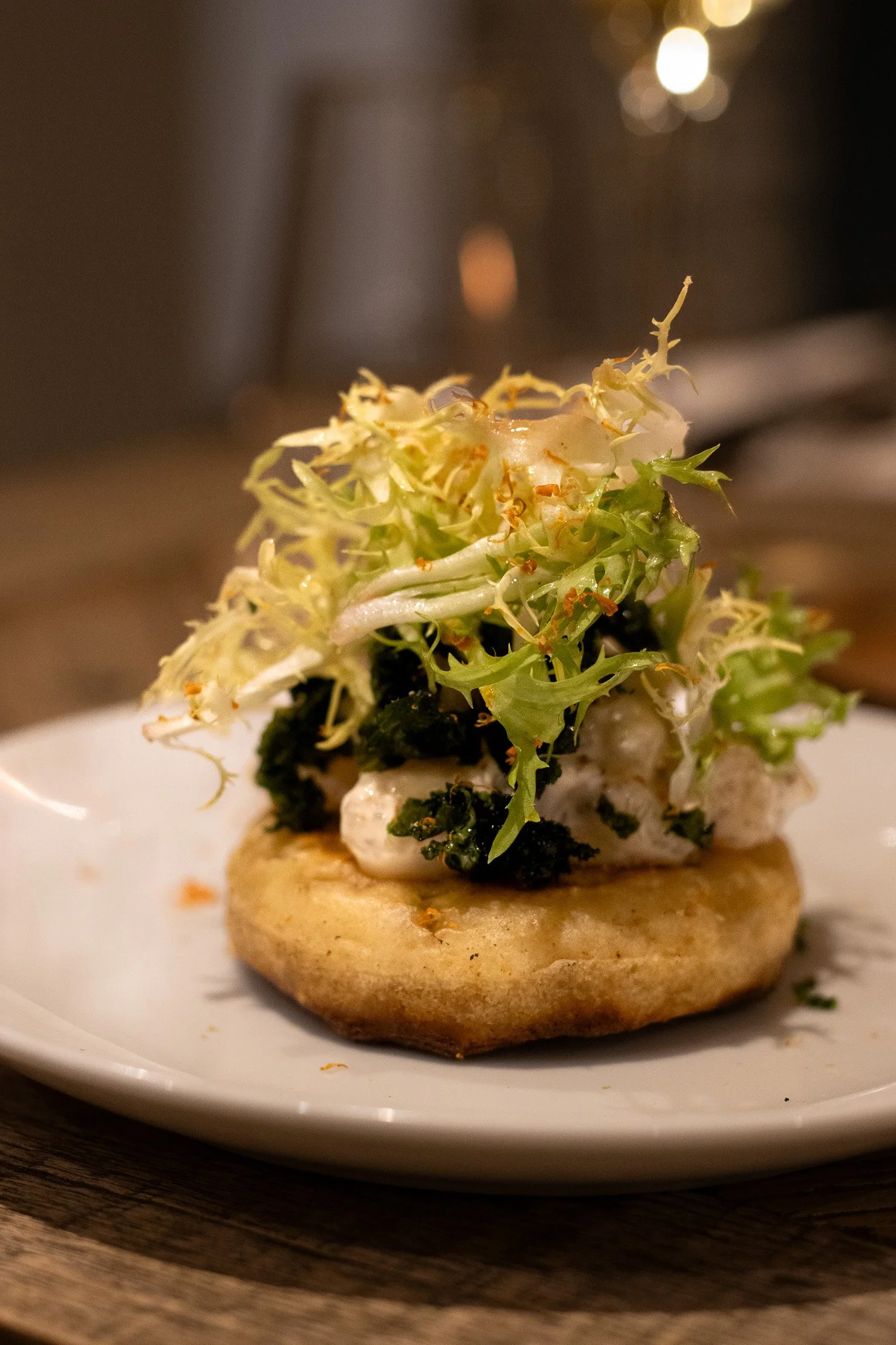 A small gourmet appetizer featuring a toasted round piece of bread topped with goat cheese, wilted greens, and microgreens, served on a white plate. The Lamb Inn. 