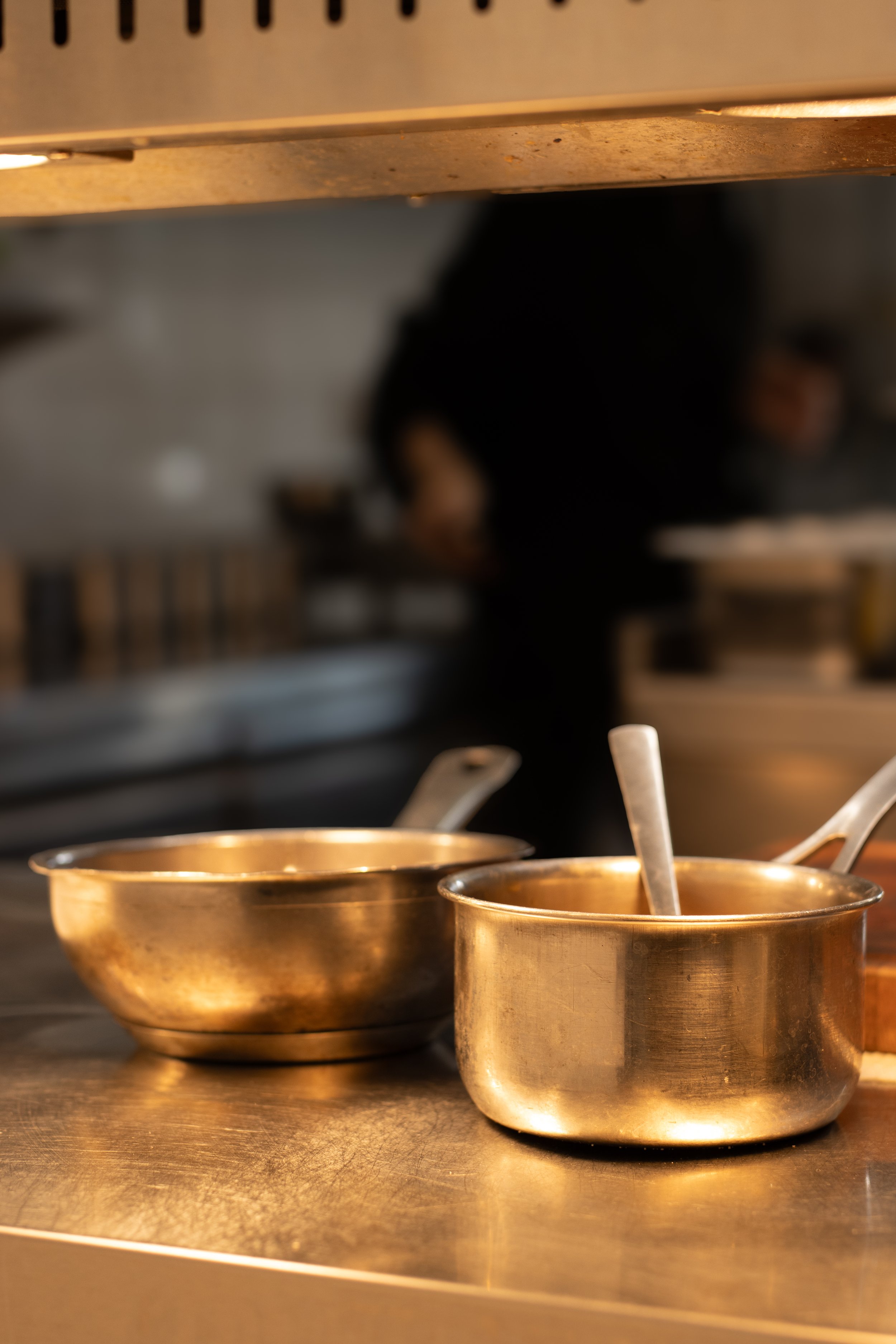 Two brass serving bowls with spoons on a stainless steel countertop in a kitchen. The Lamb Inn. 