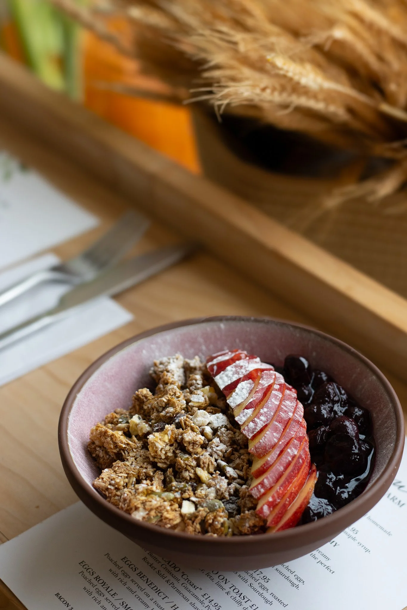 A bowl of yogurt topped with granola, sliced strawberries, and blueberries, placed on a menu or paper on a wooden table. The Cotswold Farm. 