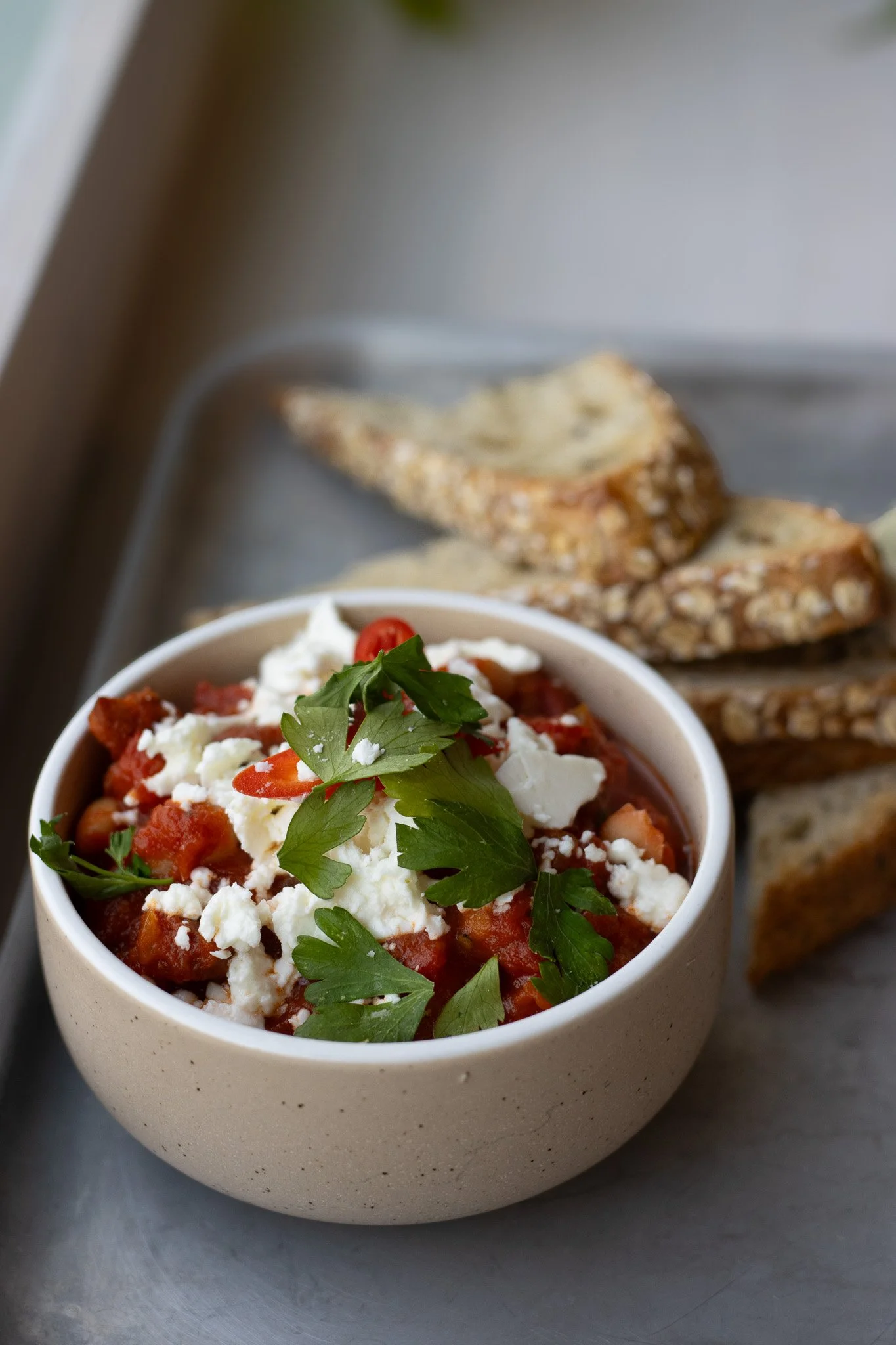 A bowl of tomato-based dish topped with crumbled cheese and fresh parsley, with bread in the background. The Cotswold Farm. 