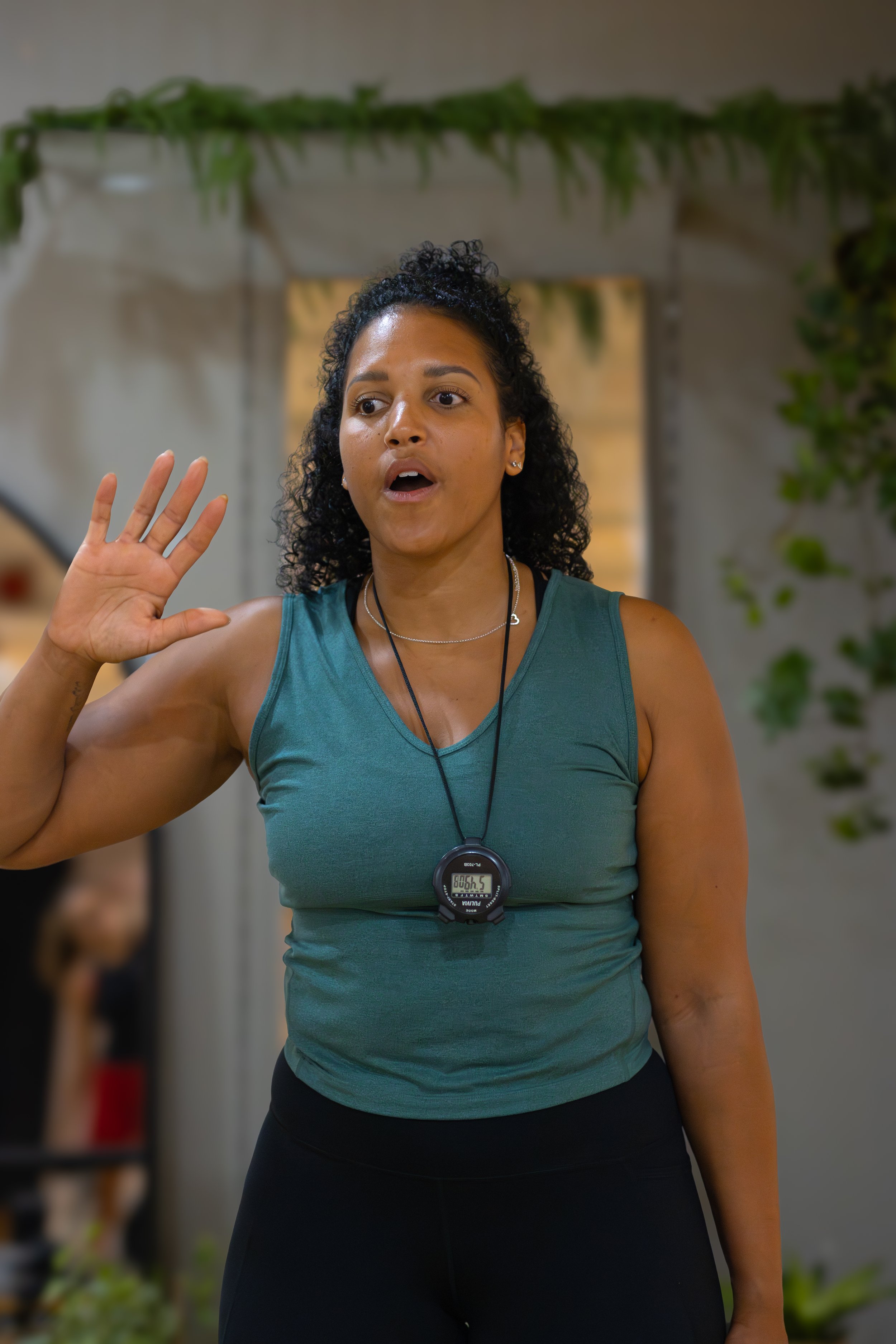 A woman in workout clothes using a stopwatch during a fitness class, with her hand raised and her mouth open. The Studio Cheltenham, Reformer Pilates. 