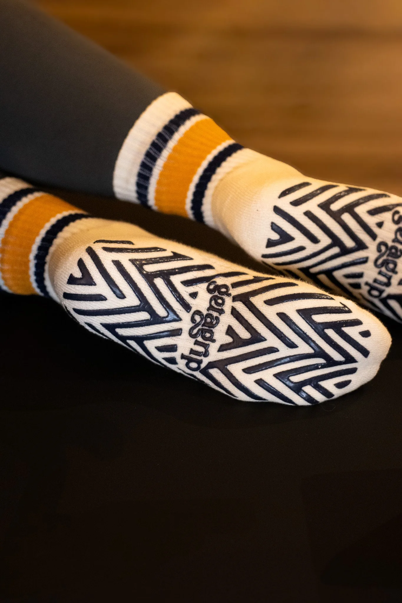 Close-up of a person's legs wearing white socks with black and yellow stripes, and text in black; the person is also wearing dark pants and sitting against a wooden surface. GetAGrip Apparel. 