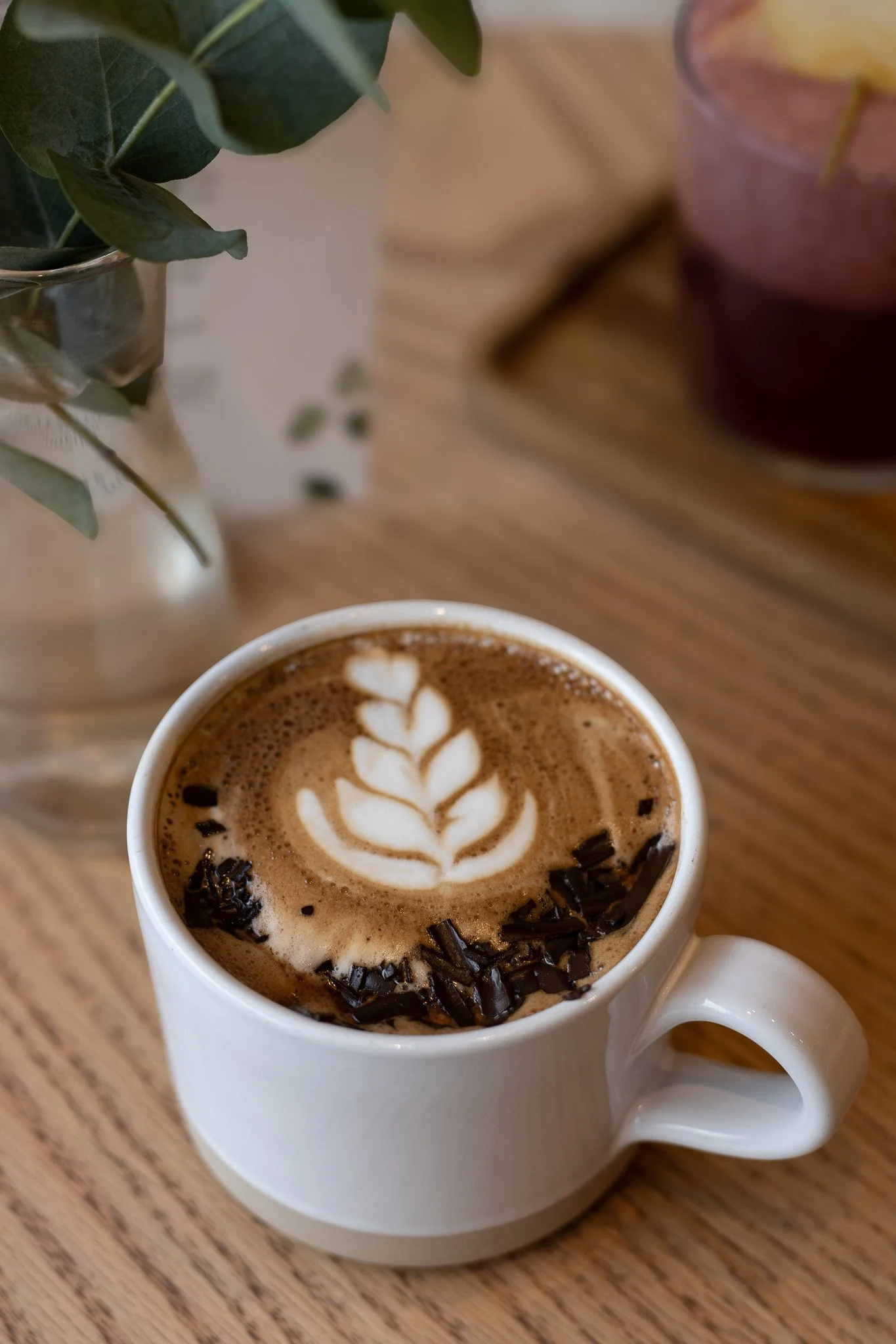 A white mug of hot chocolate with latte art and chocolate shavings, placed on a wooden table, with a vase of green leaves on the left and a pink drink on the right. The Cotswold Farm. 