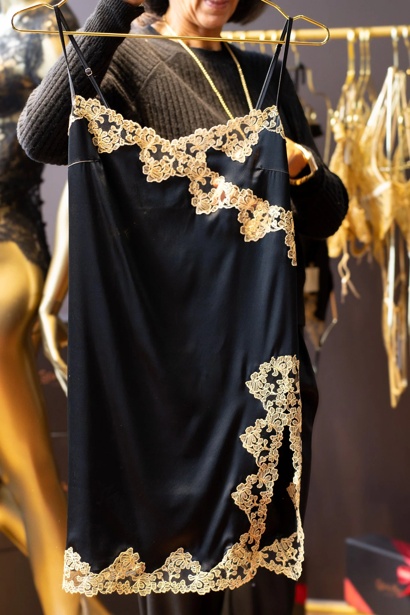 Close-up of a woman holding a black satin slip dress with gold embroidered lace detailing, hanging on a gold hanger in a clothing store. The woman is wearing a black sweater and gold jewelry. Coco De Mer.