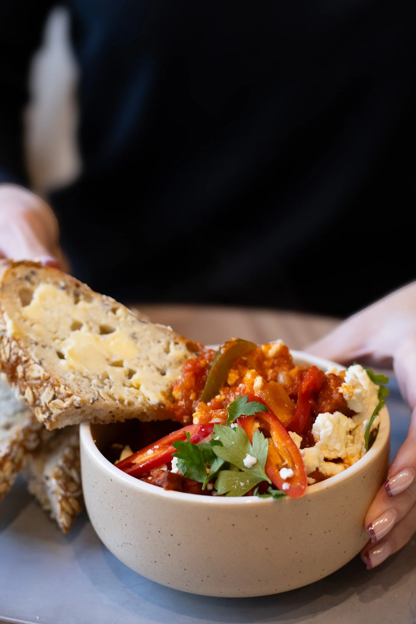 A bowl of chili topped with shredded cheese, red and green peppers, and cilantro, with a piece of bread on the side, held by a person's hand. The Cotswold Farm. 