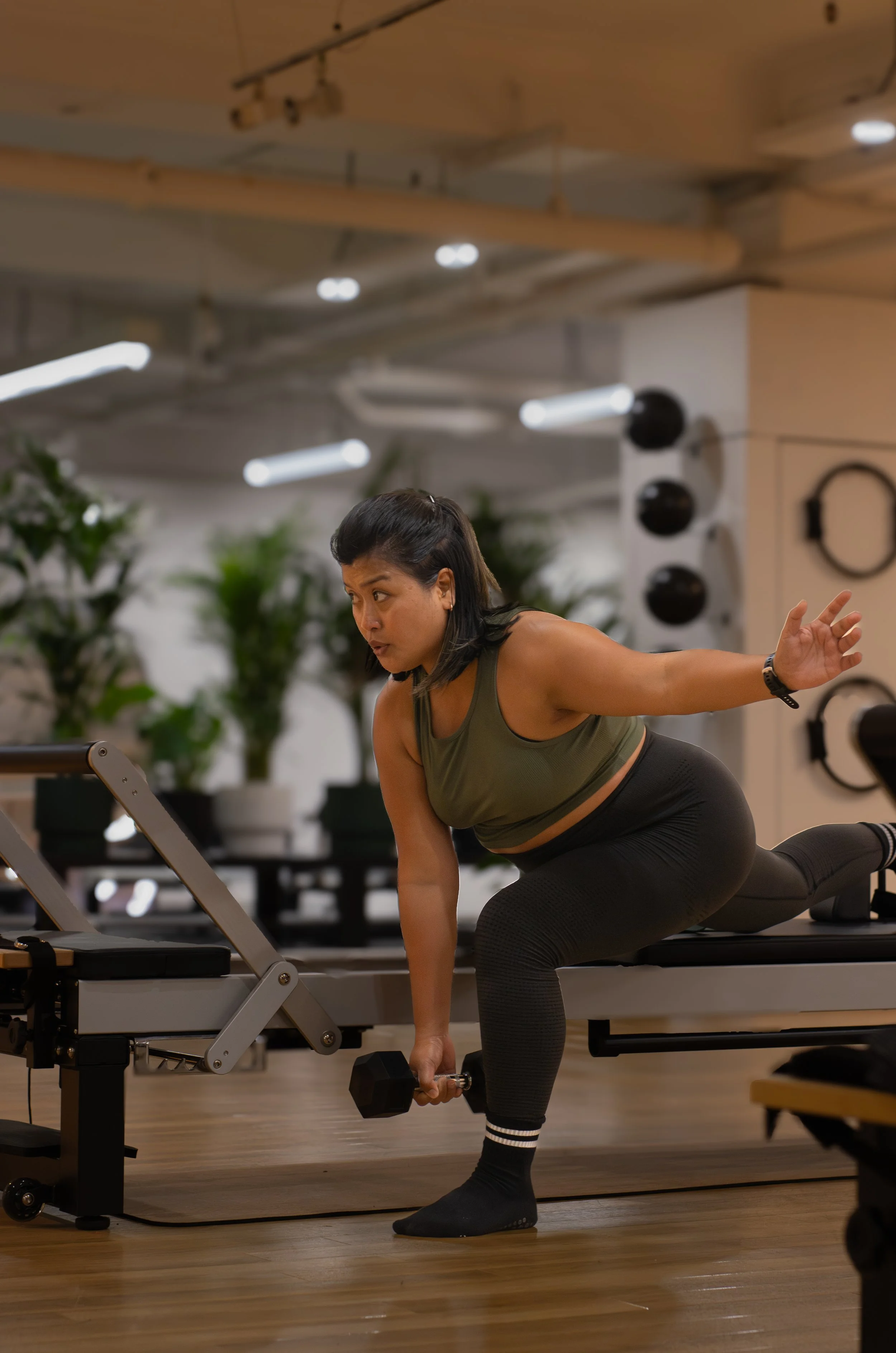 A woman with dark hair exercising in a gym. She is in a kneeling position, balancing with one hand on a bench and lifting a dumbbell with the other. The Studio Cheltenham, Reformer Pilates. 
