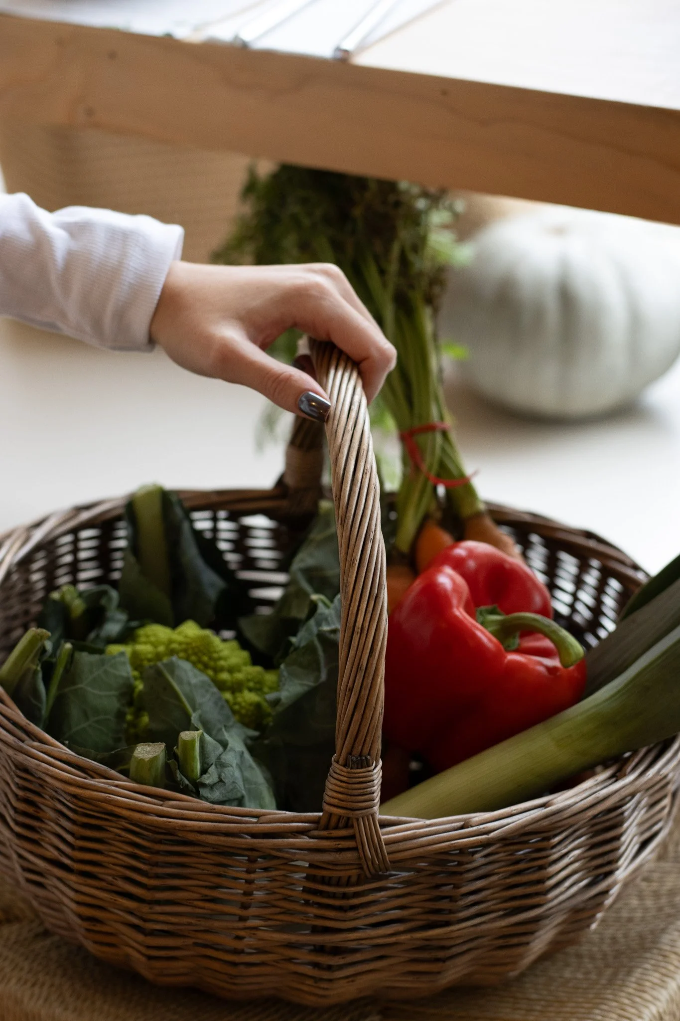 A hand reaching into a wicker basket filled with fresh vegetables, including a red bell pepper, green broccoli, and various leafy greens, with a blurred background of a wooden beam and a white bowl. The Cotswold Farm. 
