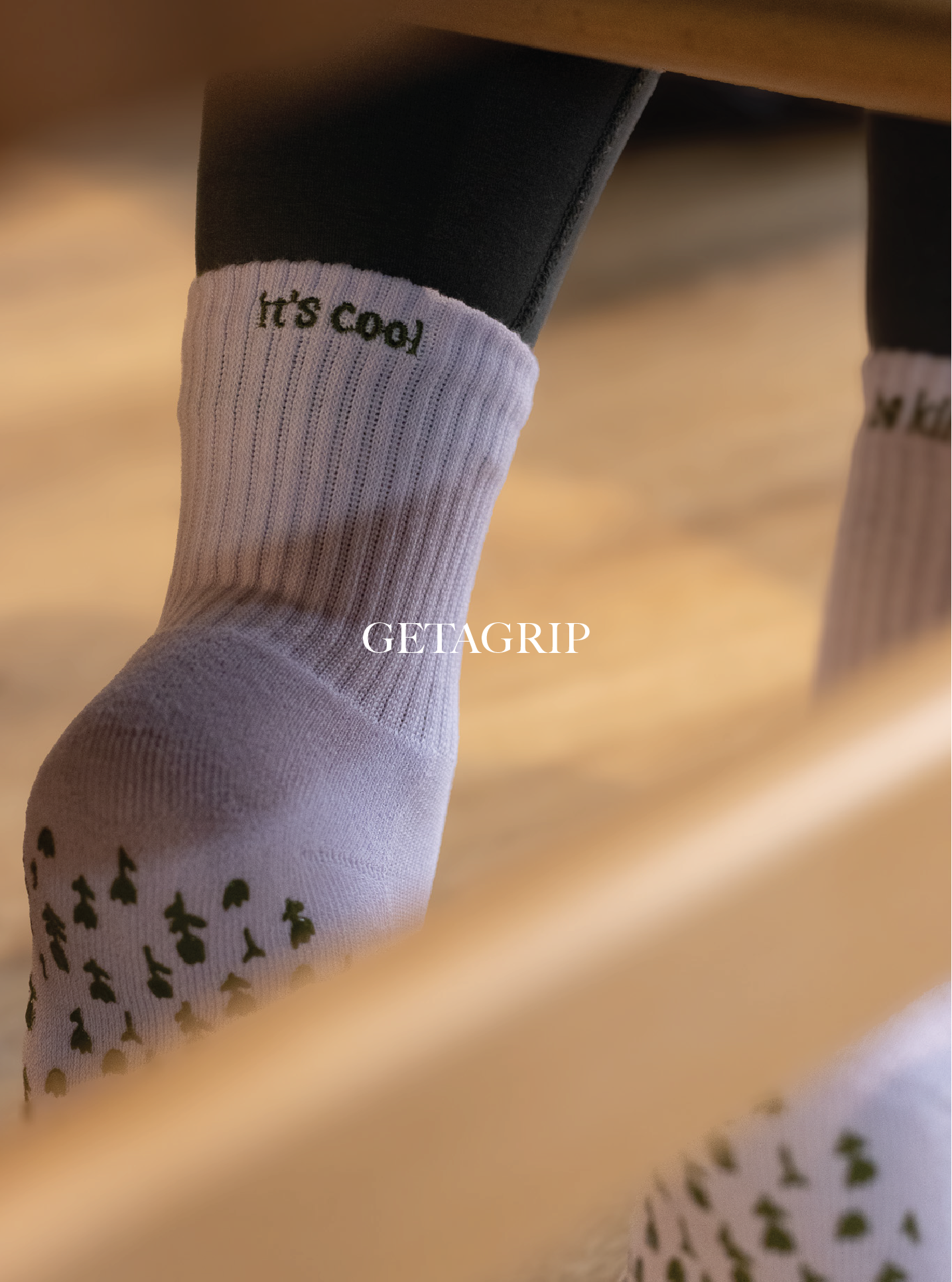 Close-up of a person's foot wearing a white sock with the text 'It's cool' embroidered near the ankle, and a black shoe. The background is blurred with warm tones. Get A Grip Apparel.
