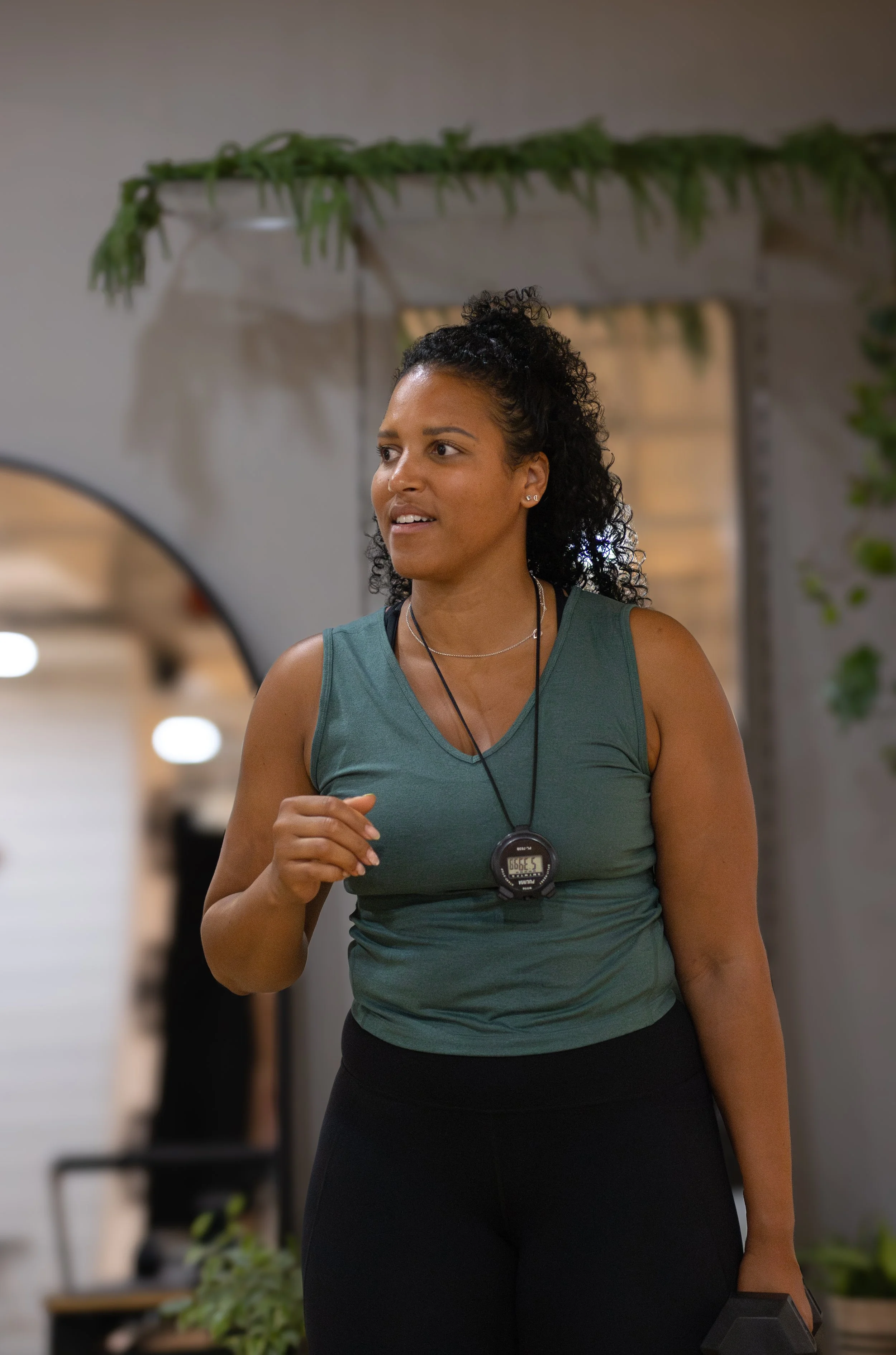 A woman in a green workout tank top and black leggings holding a black dumbbell in her right hand, wearing a fitness tracker or stopwatch around her neck. The Studio Cheltenham, Reformer Pilates. 