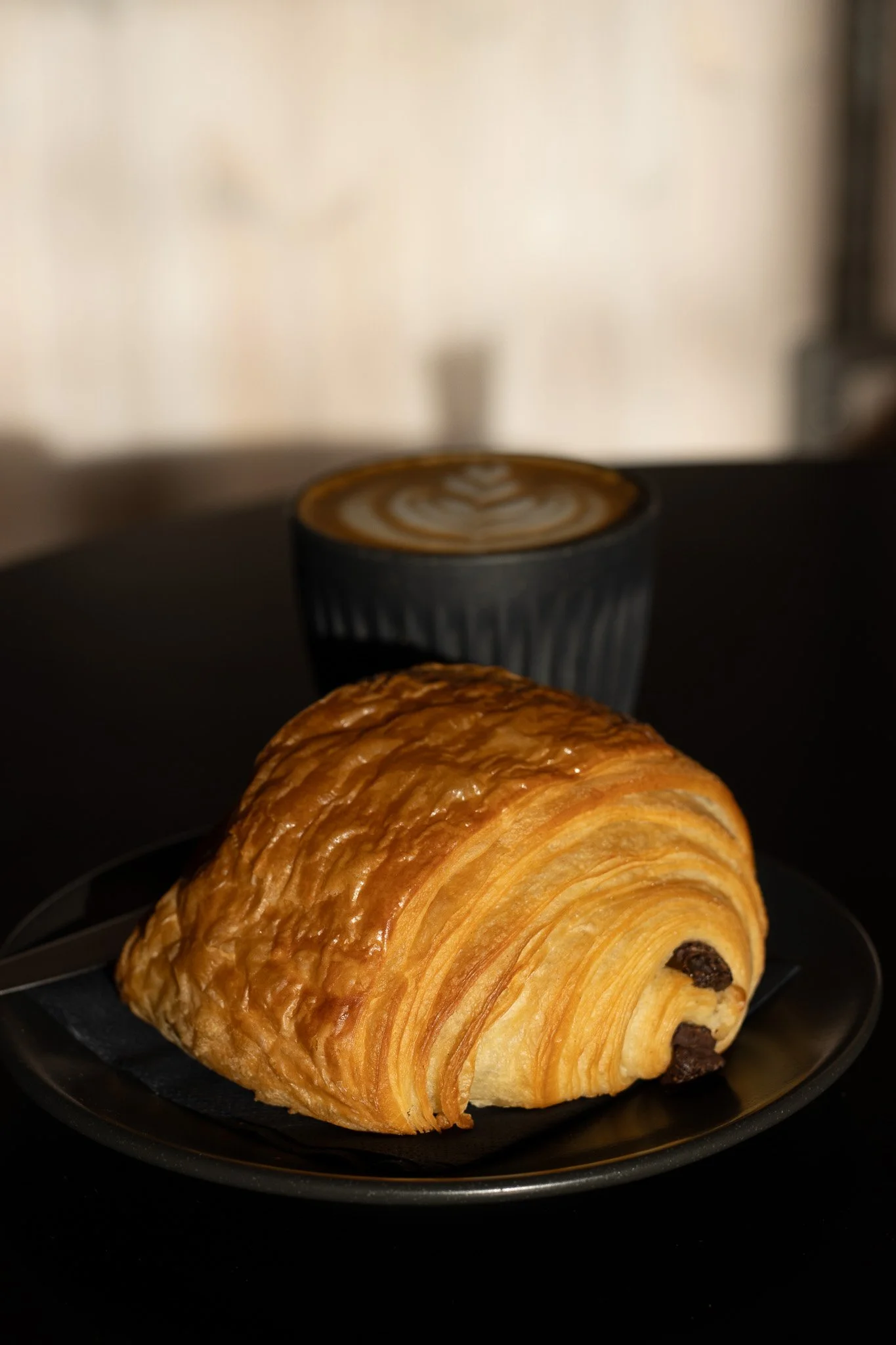 A Pain Au Chocolat croissant with chocolate chips on a black plate in front of a blurred background with a cup of latte art coffee. The Scandinavian Coffee Pod.