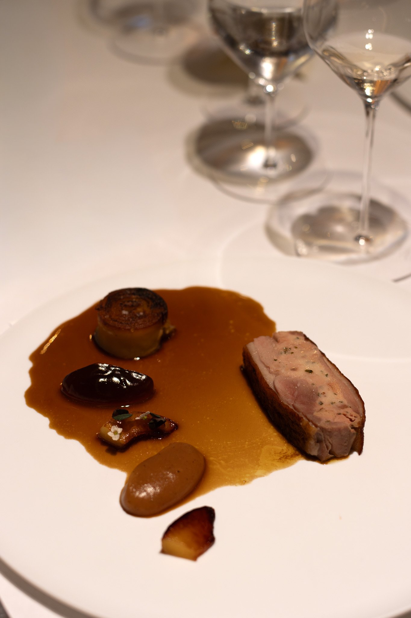 Close-up of gourmet appetizer featuring sliced meat, sauces, and garnishes on a white plate, with empty wine glasses in the background. Petrus.