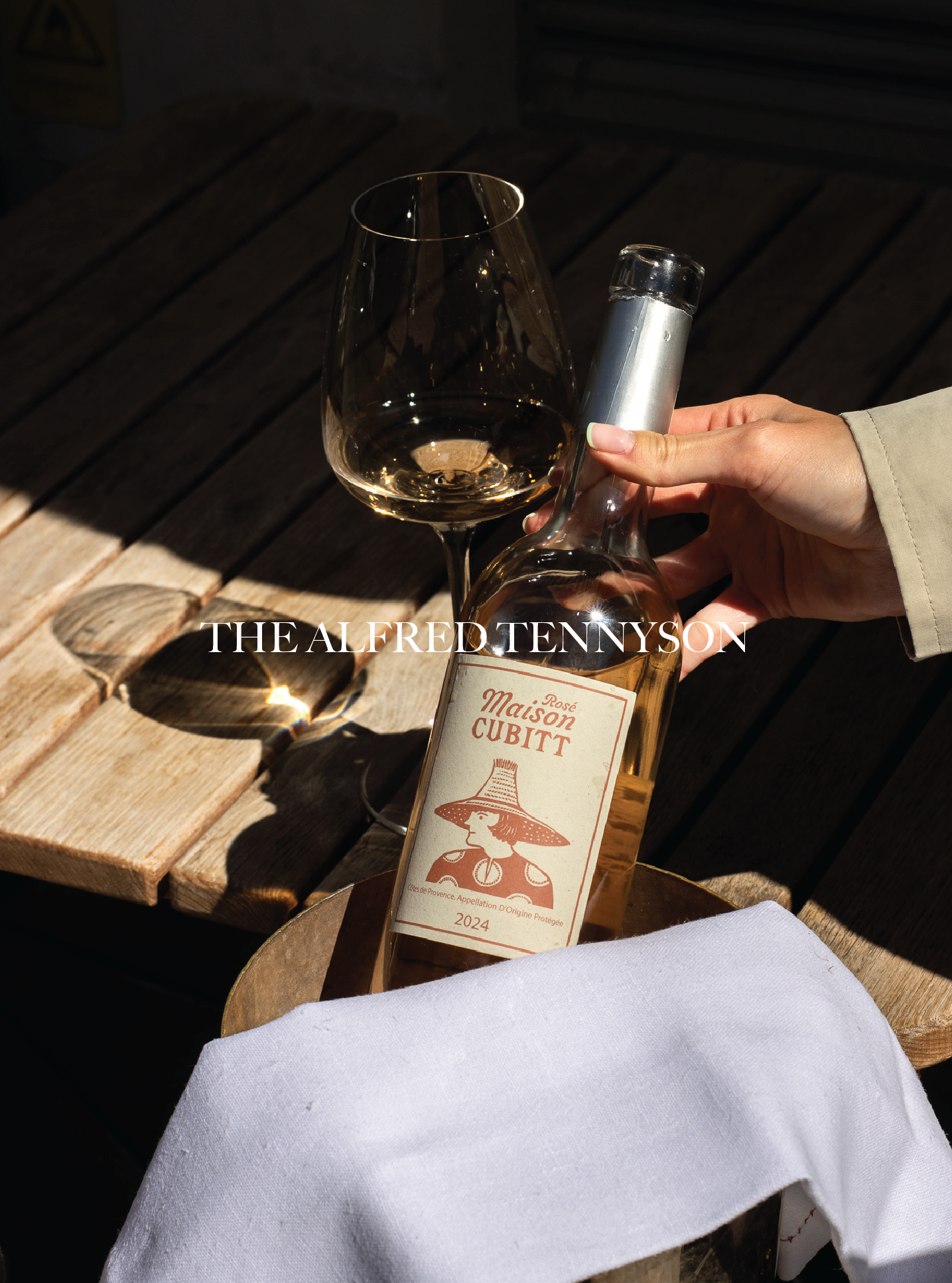 A hand holding a wine bottle labeled 'Maison Cubitt Rosé 2024' next to a glass of white wine on a wooden table with a cloth underneath. The Alfred Tennyson.