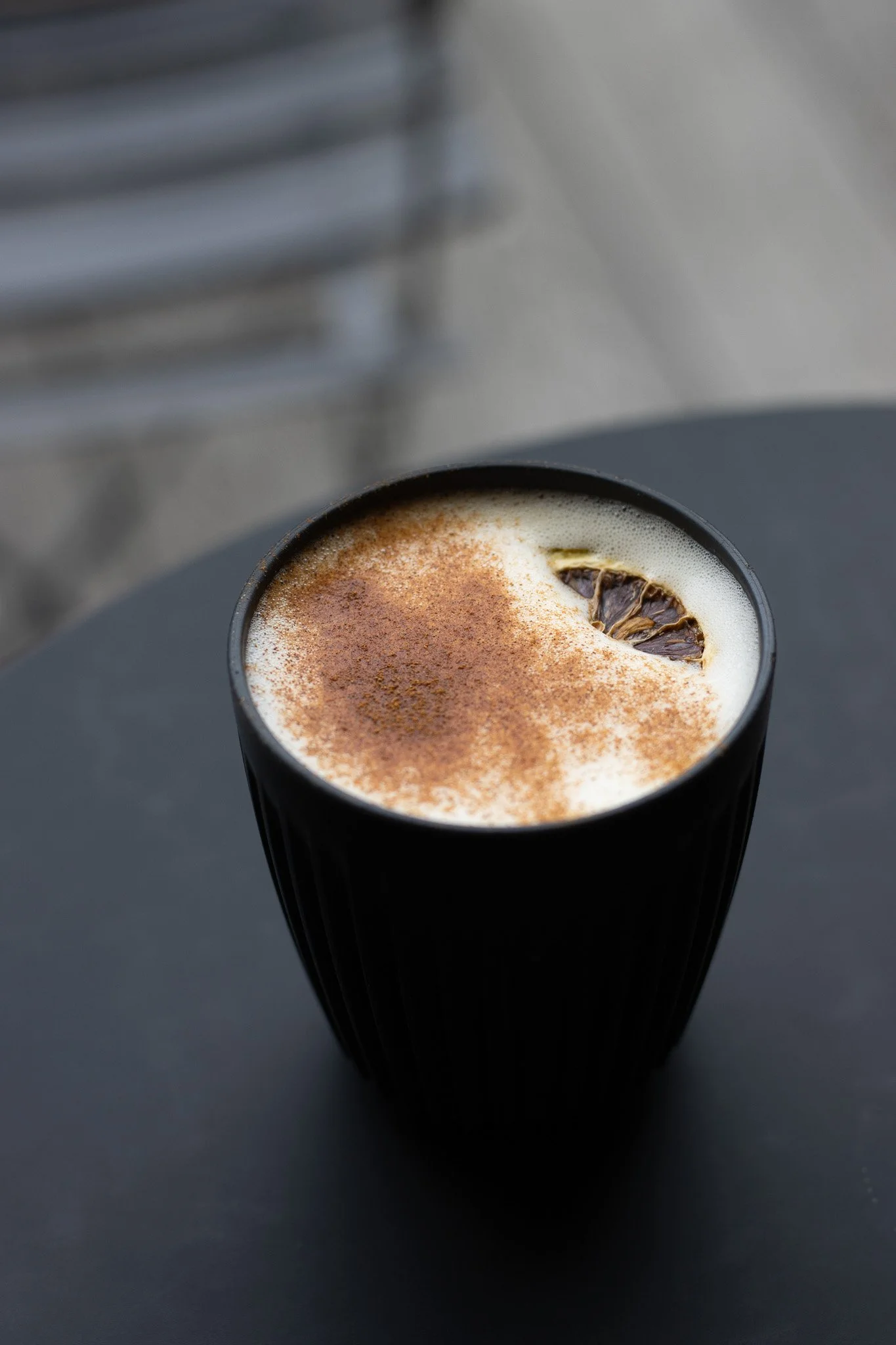 Close-up of a black cup filled with frothy coffee, topped with a sprinkle of cinnamon and garnished with a dried lemon slice, placed on a dark surface. The Scandinavian Coffee Pod.