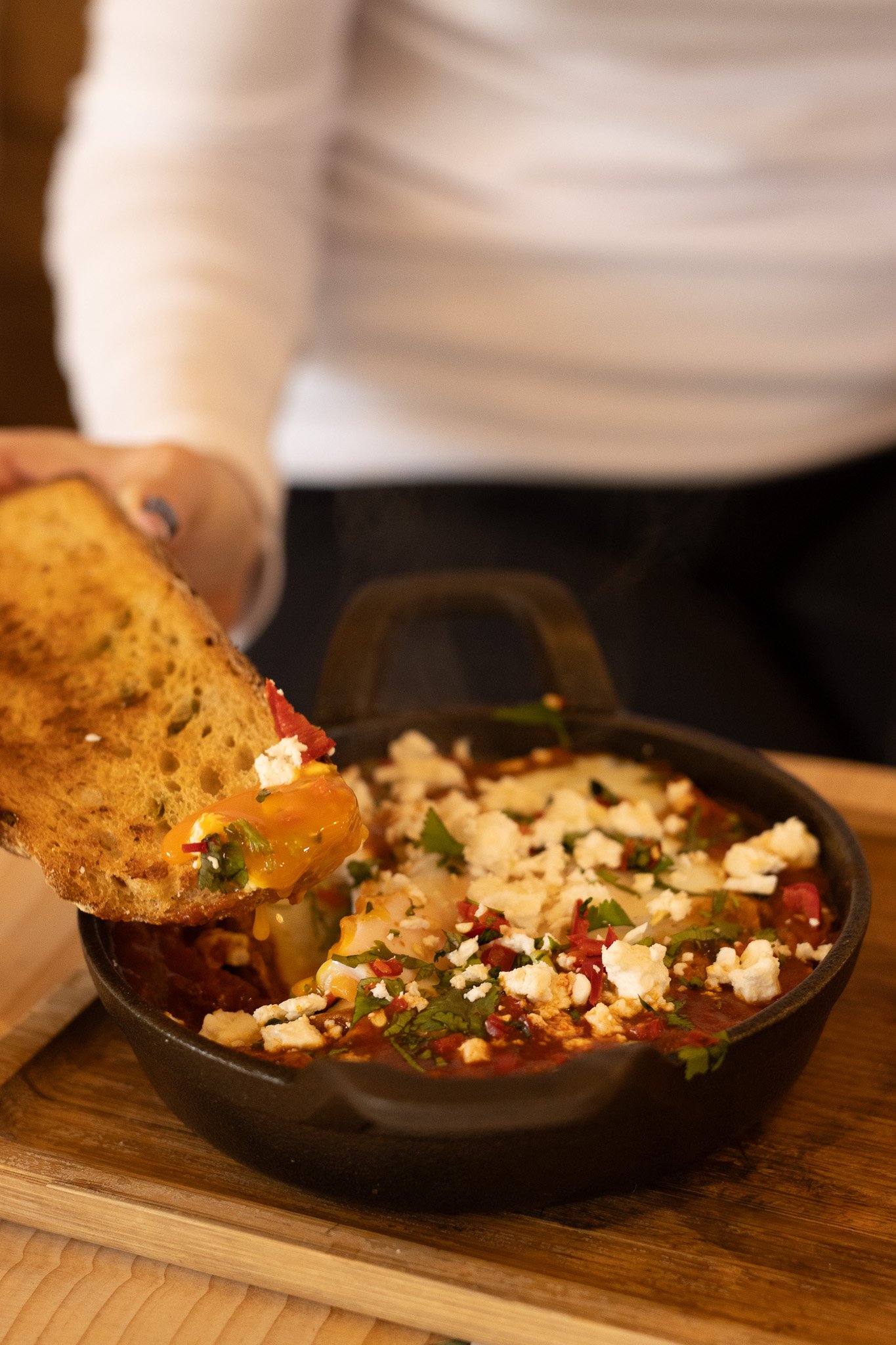 Close-up of a hand holding a piece of bread with cheese, hot sauce, and herbs. In the background, a bowl of Mexican food with cheese, cilantro, and red sauce on a wooden tray. The Cotswold Farm. 