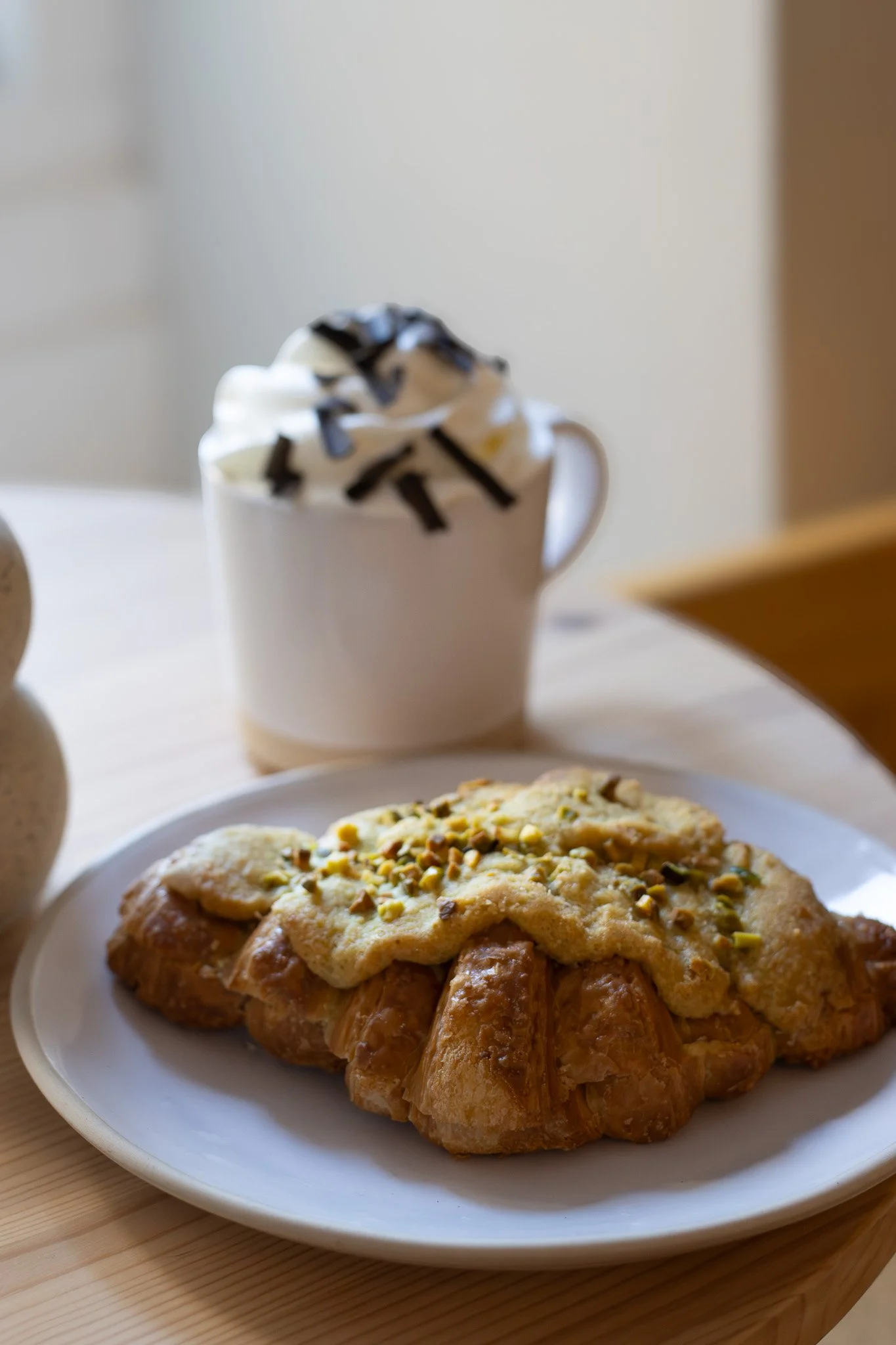 A white plate with a croissant topped with glazed cream and chopped pistachios. In the background, a cup of whipped cream with chocolate drizzle and a dessert, possibly a cookie or cake, on a white plate. The scene is set on a light wooden surface. 