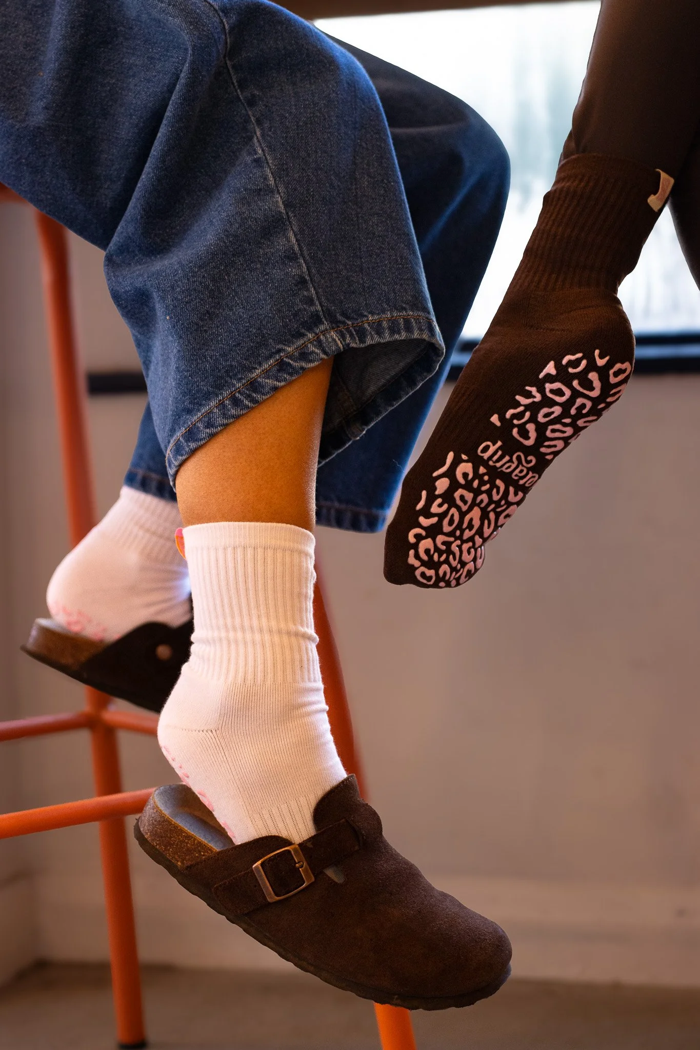Close-up of a person wearing dark brown socks with pink text, white knee-high socks, black shoes with a buckle, denim shorts, and a black sweater, sitting on an orange chair. GetAGrip Apparel. 