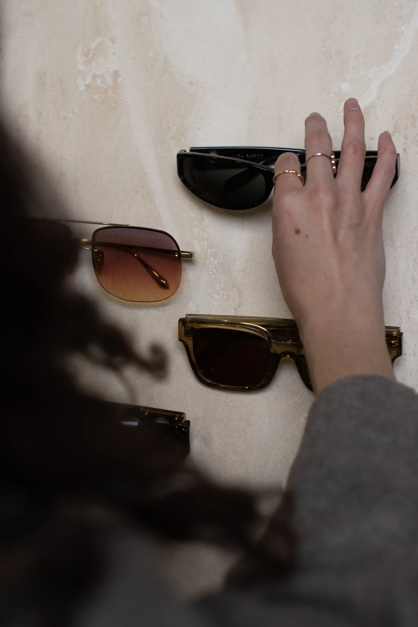 A person with rings on their fingers adjusting black sunglasses on a beige surface, with other sunglasses in different styles and colors nearby. Linda Farrow.