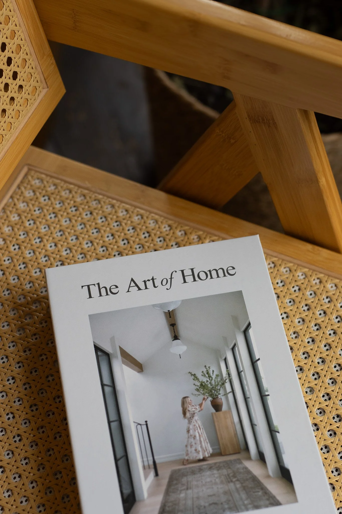 Cover of a book titled "The Art of Home" showing an interior space with tall windows, a woman in a floral dress arranging a plant on a wooden stand, and minimalist decor. The New England Coffee House.