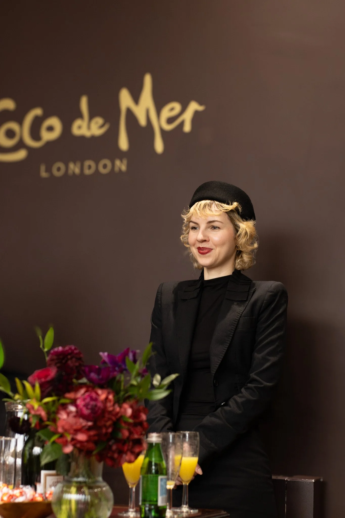 A woman with blonde curly hair and red lipstick wearing a black beret and black blazer standing behind a table with a bouquet of flowers, glasses of orange juice, and bottles, in front of a brown wall with gold text that reads 'Coco de Mer London.'
