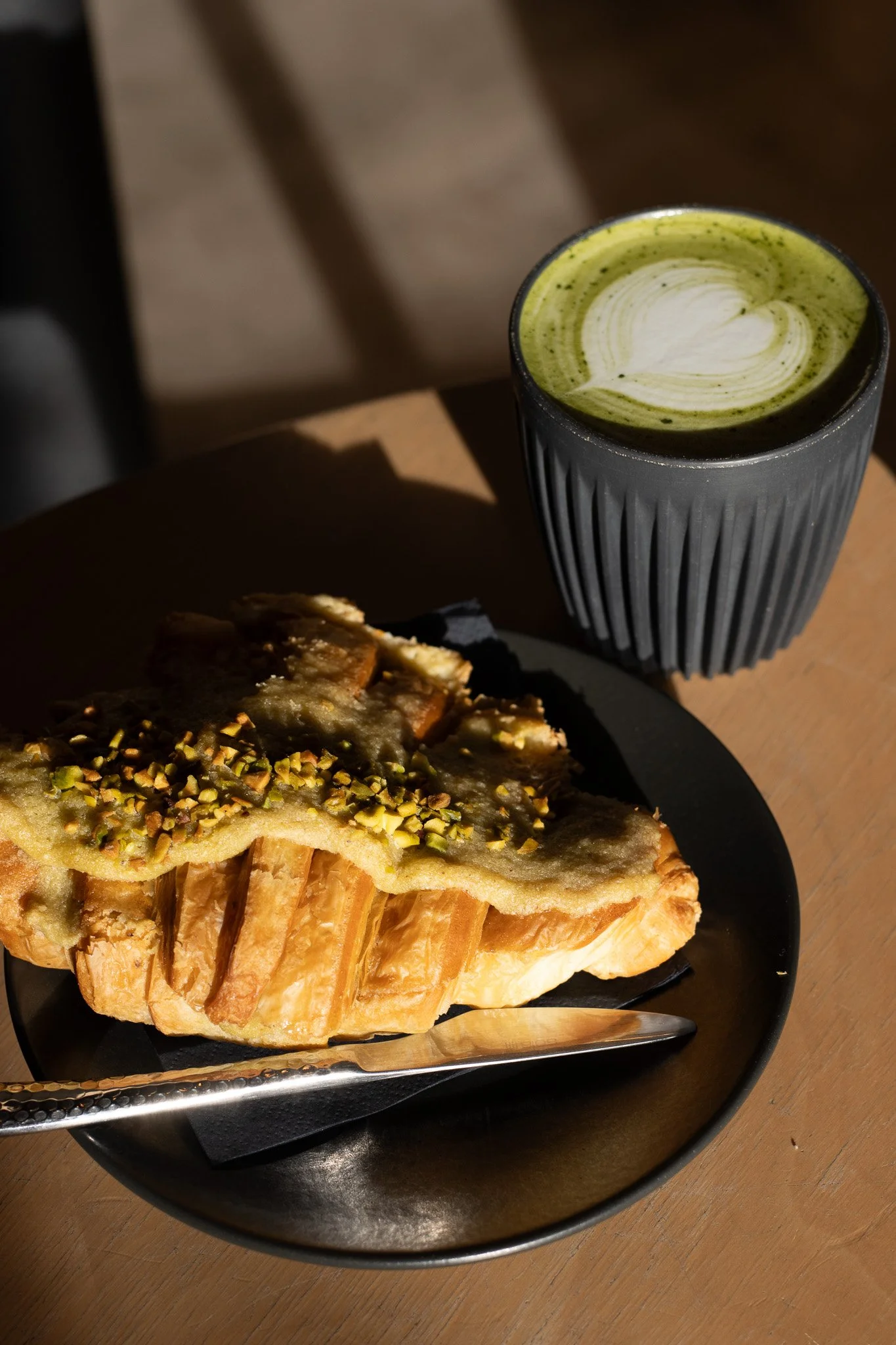 A black plate with a layered pastry topped with chopped pistachios and a slice of roasted vegetable. In the background, a black ribbed ceramic cup with green matcha latte with latte art and a heart shape on top, placed on a wooden table. The Scandina