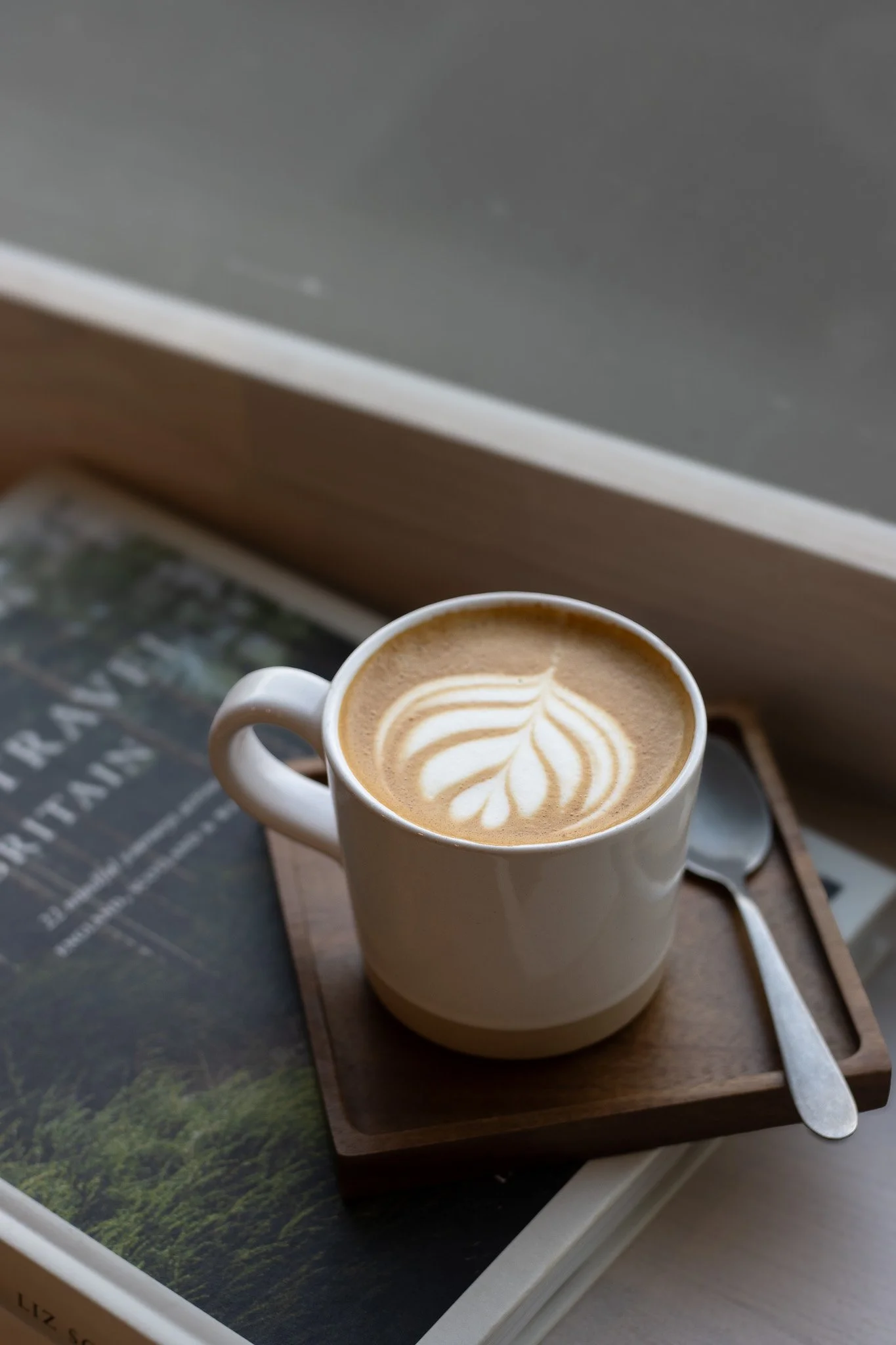 A white mug of coffee with latte art sitting on a wooden tray on a table near a window. The Cotswold Farm. 