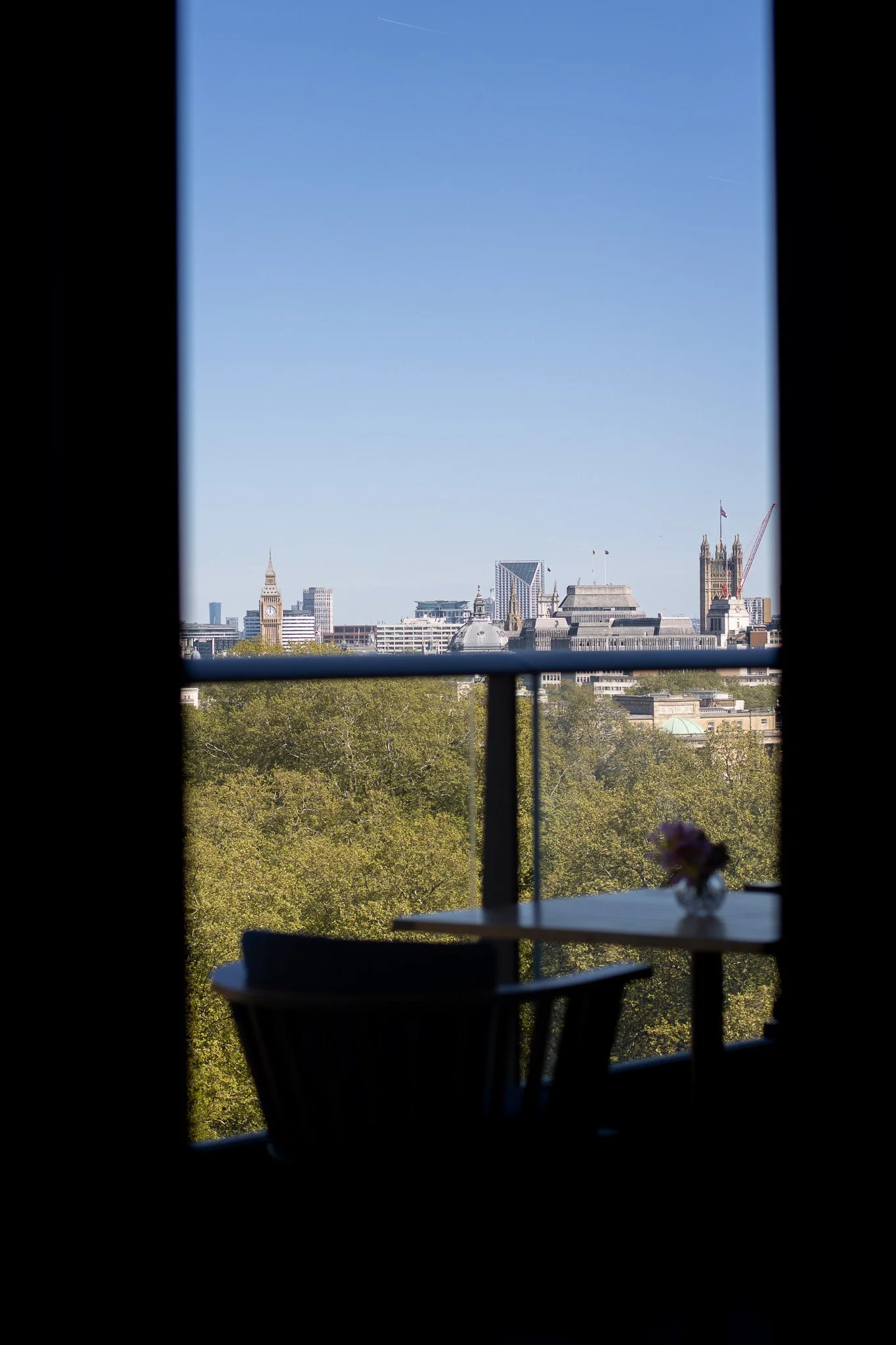A view of a city skyline through a window, featuring famous landmarks such as Big Ben and modern buildings, with greenery in the foreground and the sky in the background. The Peninsula.