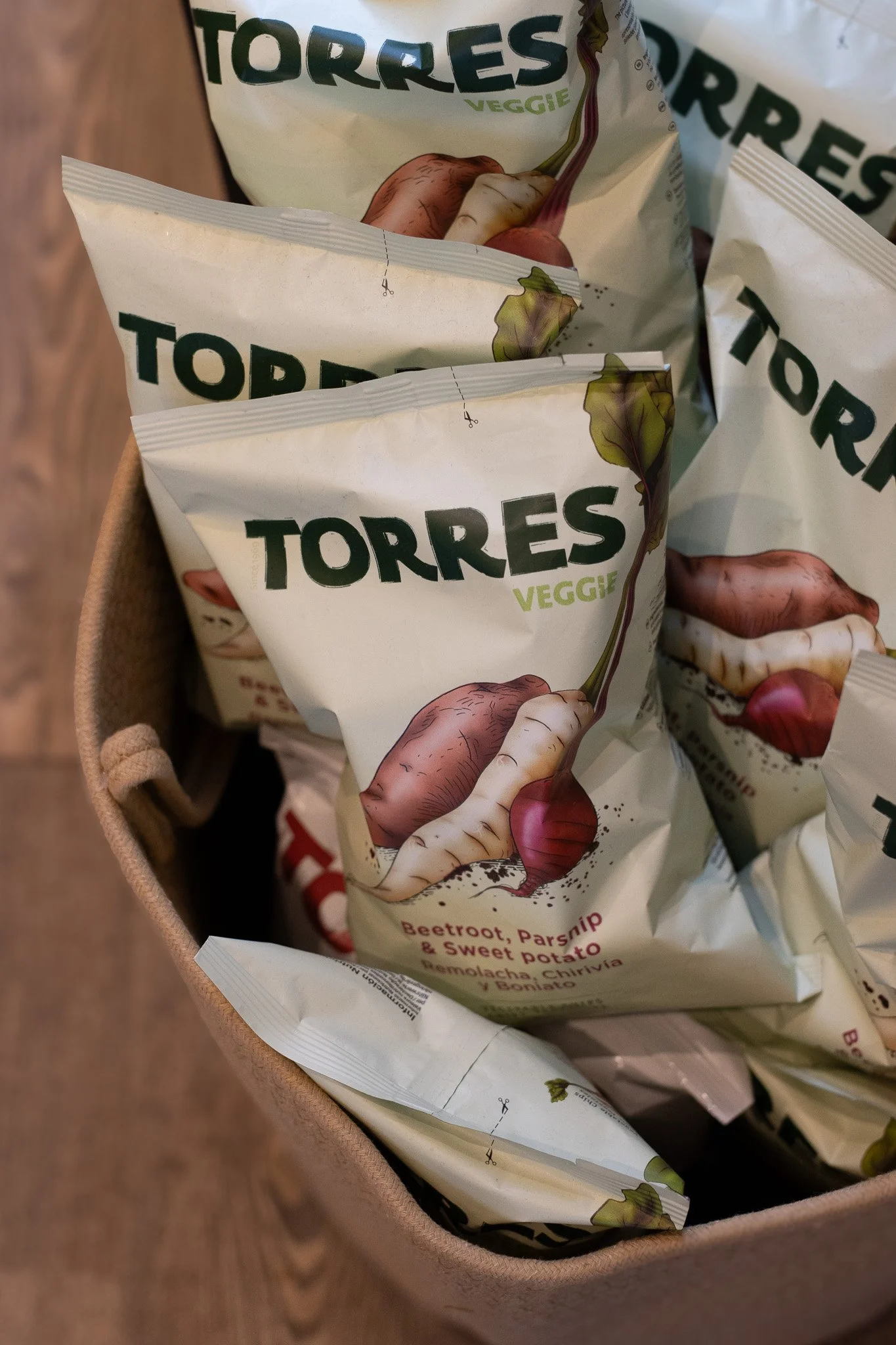 Close-up of grocery bags labeled 'Torres Veggie' with illustrations of vegetables and text indicating ingredients like beetroot, parsnip, and sweet potato, placed inside a beige basket. The Cotswold Farm. 