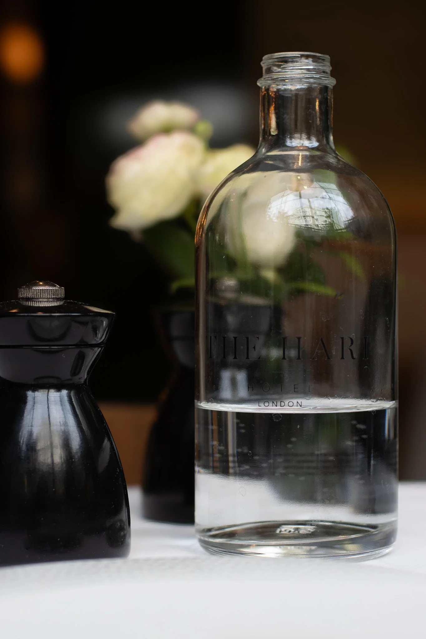 A clear glass bottle with the words 'The Hari Hotel London' etched on it, surrounded by black ceramic containers with silver lids, and blurred flowers in the background. The Hari, Il Pampero.