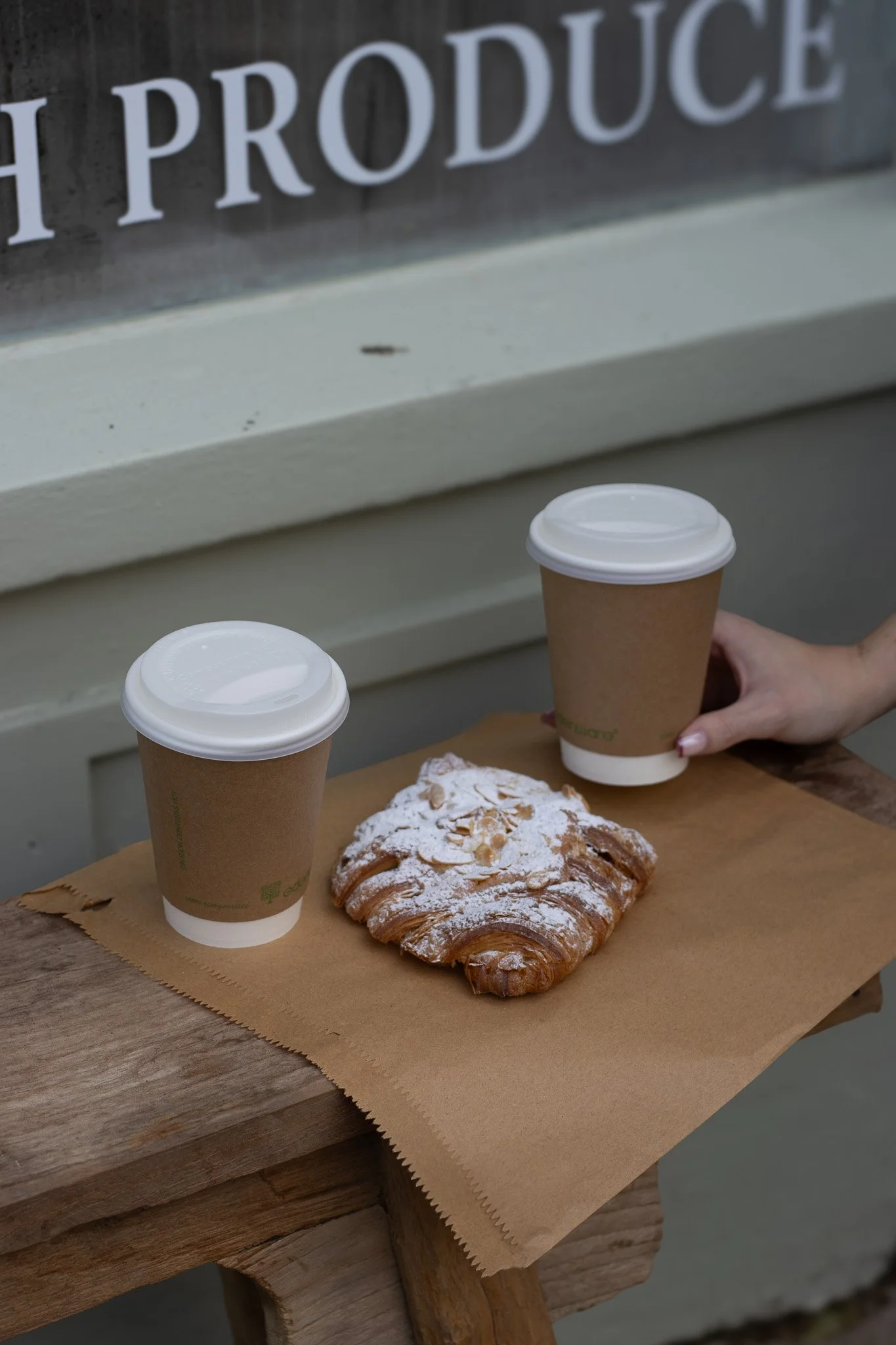 Two disposable coffee cups with white lids on a wooden table, with a pastry dusted with powdered sugar and sliced almonds in front of them, and a blurry sign in the background reading 'The Produce'. The Cotswold Farm. 