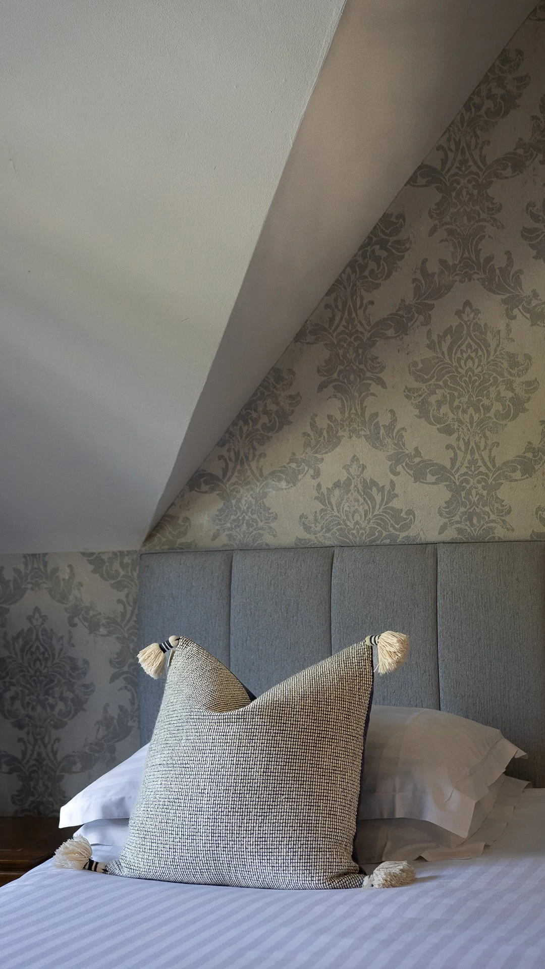 A bed with a decorative cushion and pillow, against a padded headboard, with a wall featuring patterned wallpaper and a section of a ceiling. The Lamb Inn. 