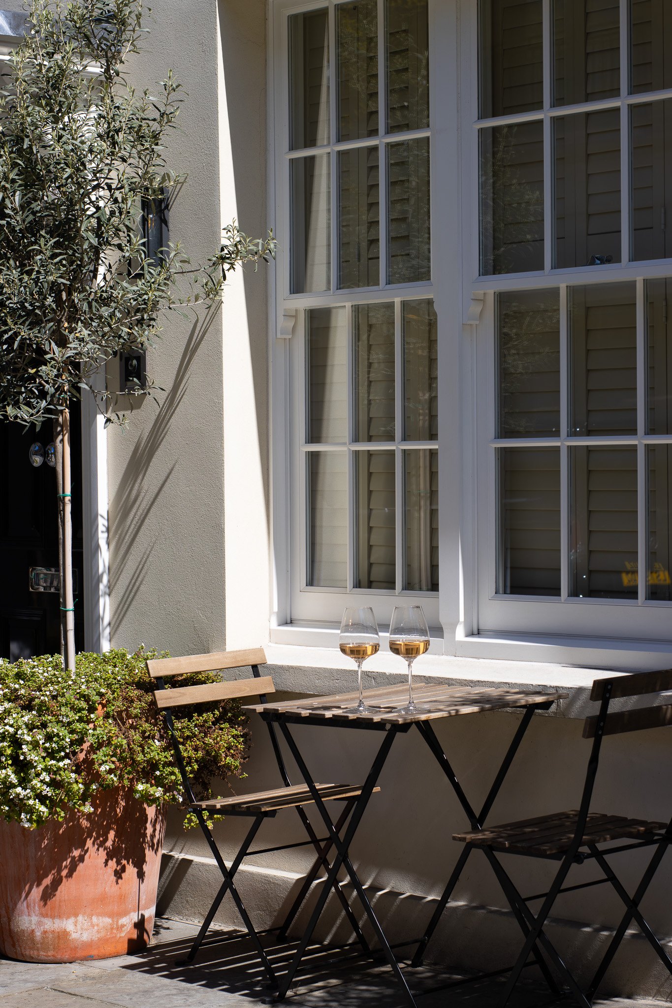 Outdoor patio area with a round wooden table, two wine glasses filled with white wine, two wooden chairs, a large potted plant with white flowers, a window with closed blinds, and part of a white building exterior bathed in sunlight. Alfred Tennyson.