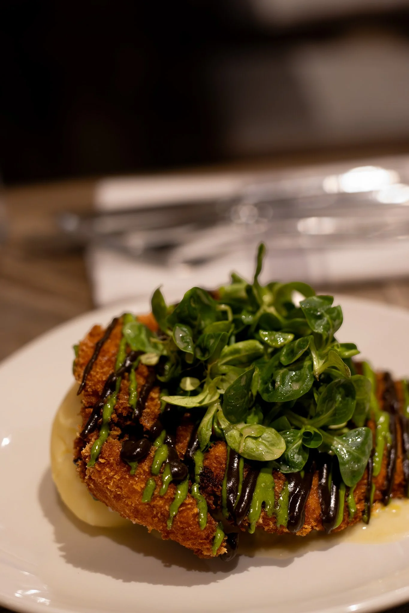 A plate of food featuring a breaded and fried meat cutlet topped with fresh green leafy salad and a drizzle of dark and green sauce, with a soft-boiled egg underneath, and a blurred background of kitchen appliances. The Lamb Inn. 