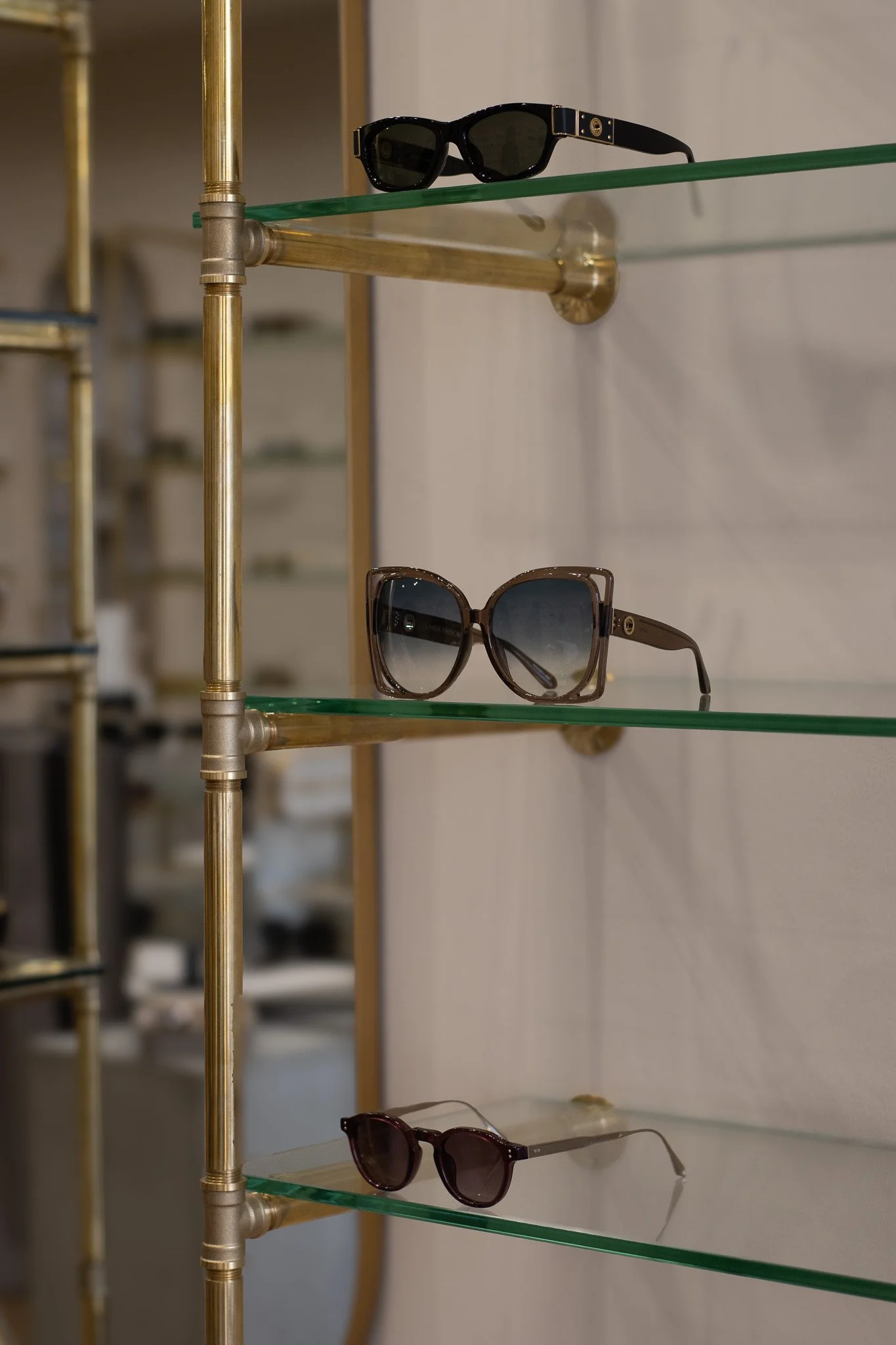 Three pairs of sunglasses on a glass and brass display shelf, with the top pair being black and the bottom pair brown, and a faded background. Linda Farrow.