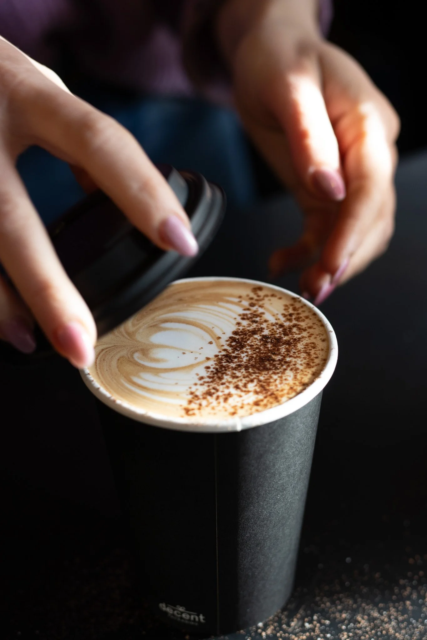 Close-up of a hand holding a black container pouring coffee onto a cup of latte with foam and chocolate sprinkles. The Scandinavian Coffee Pod.