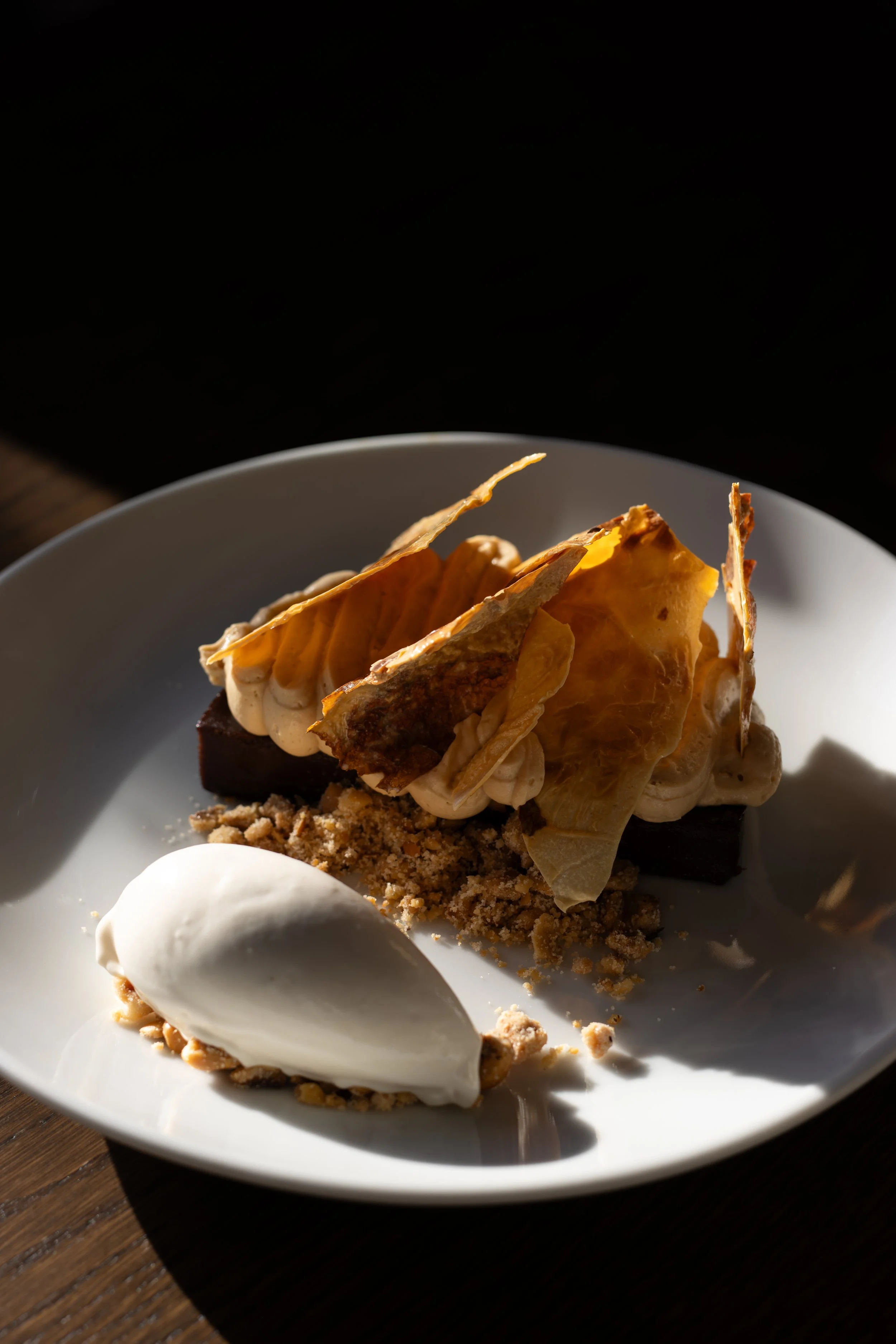 A slice of chocolate and caramel dessert on a white plate, topped with caramelized sugar shards and surrounded by crumbled biscuit and a dollop of whipped cream. The Lamb Inn. 