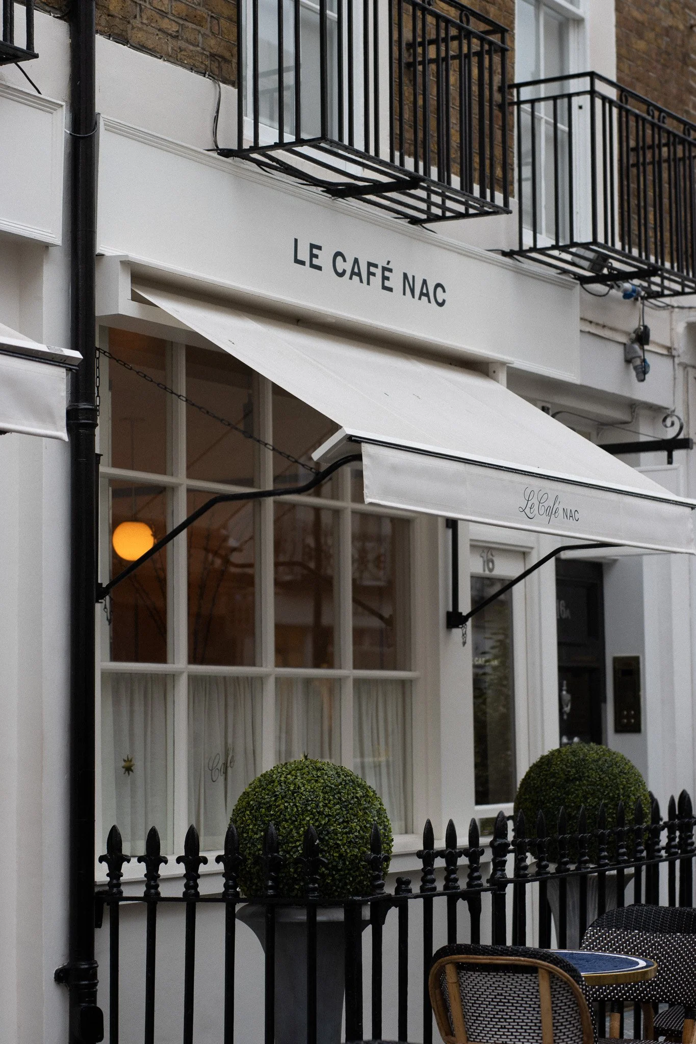 Exterior of a cafe named 'Le Café NAC' with white awning, black metal railings, glass windows, and small round bushes in front. Le Cafe NAC.