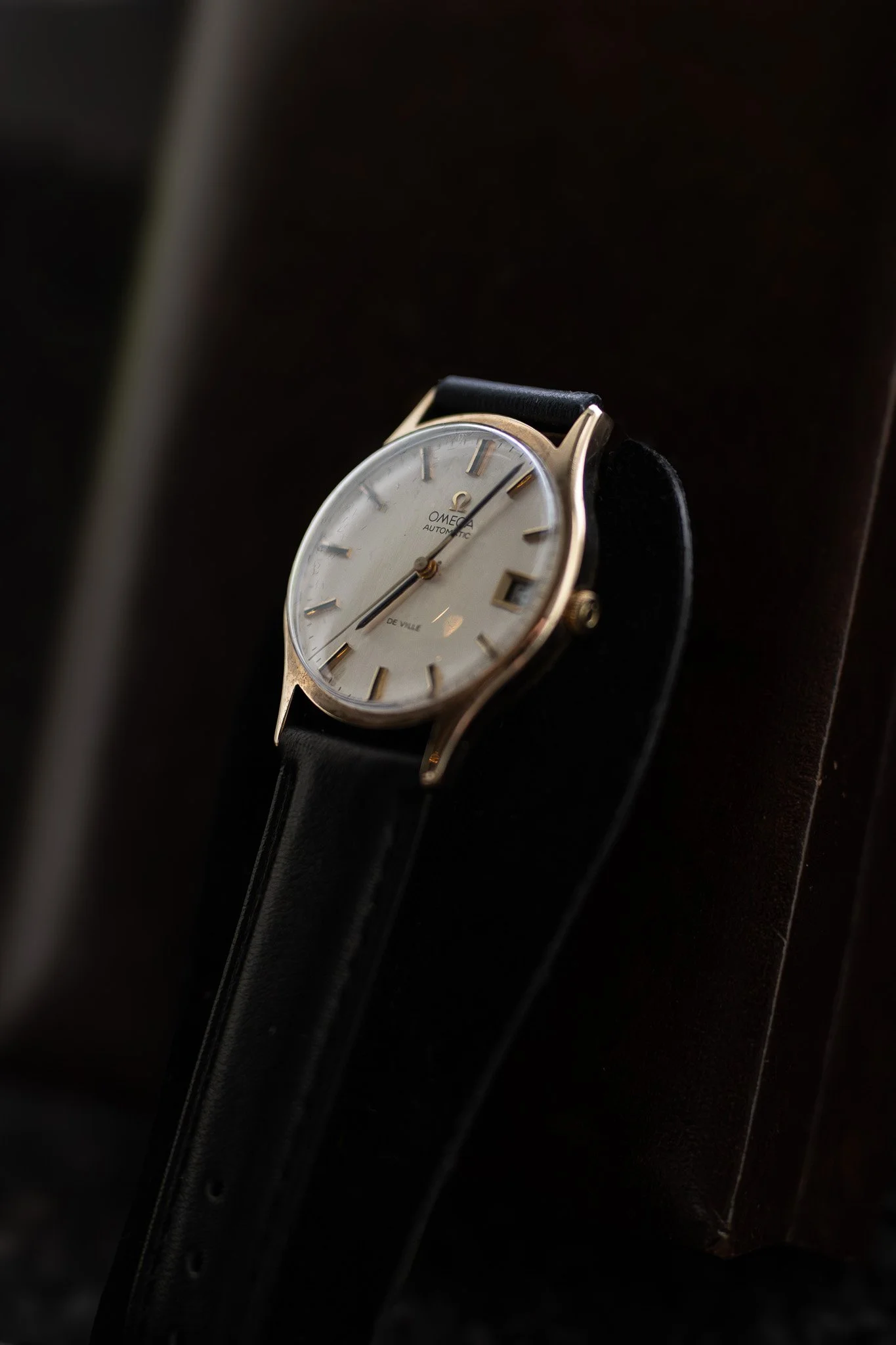 Close-up of a vintage Omega automatic wristwatch with a cream dial, gold markers and hands, and a black leather strap, resting on a dark surface. Aurmont. Watches.